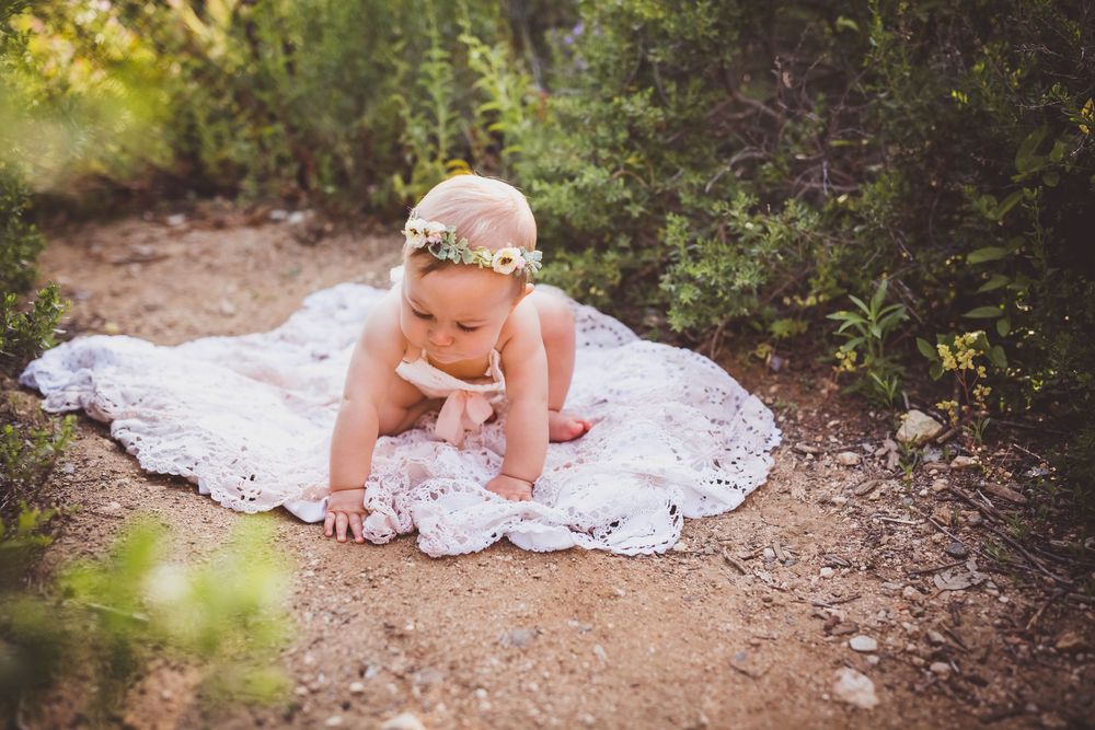 Baby girl in floral headband crawling on white lace blanket outdoors on dirt path surrounded by greenery near redlands ca