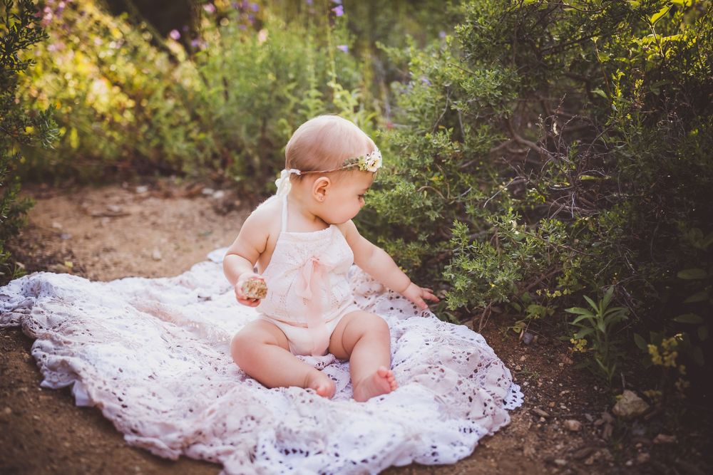 A baby girl in a white lace romper sits on a blanket outdoors surrounded by lush green plants in the inland empire, ca