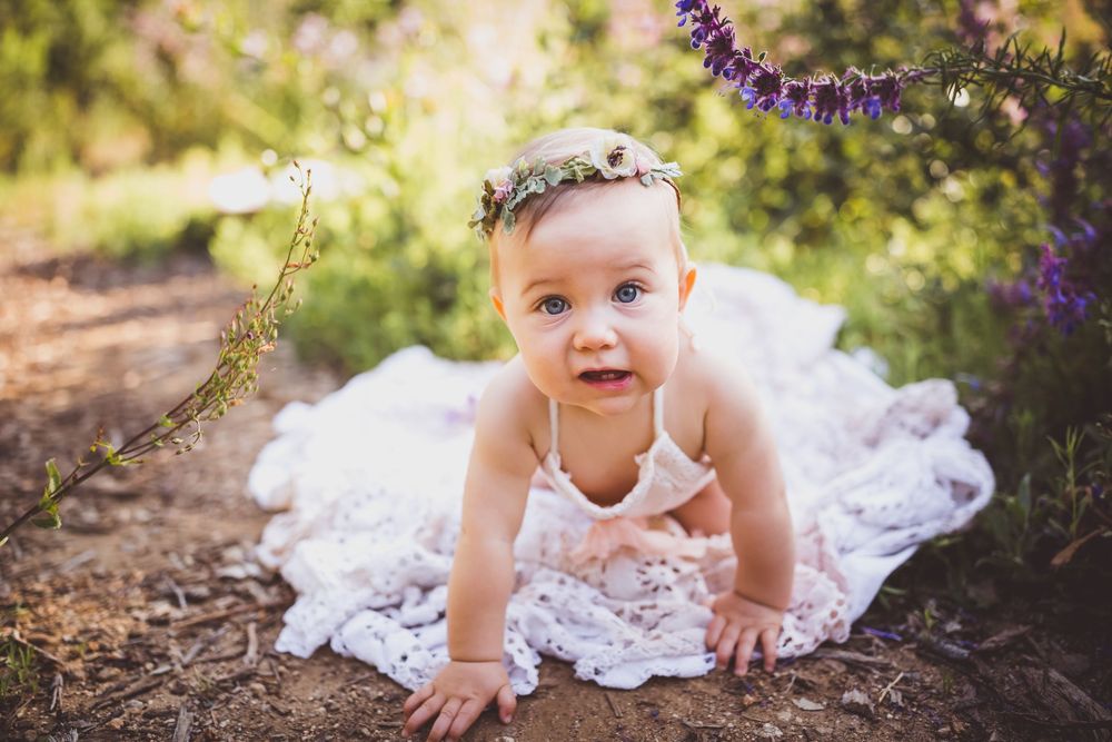 Adorable baby girl in white lace dress and floral crown crawling outdoors among purple wildflowers with heidi grace photography