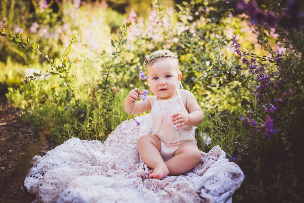 Adorable baby girl in white romper sitting on lace blanket surrounded by purple wildflowers outdoors.