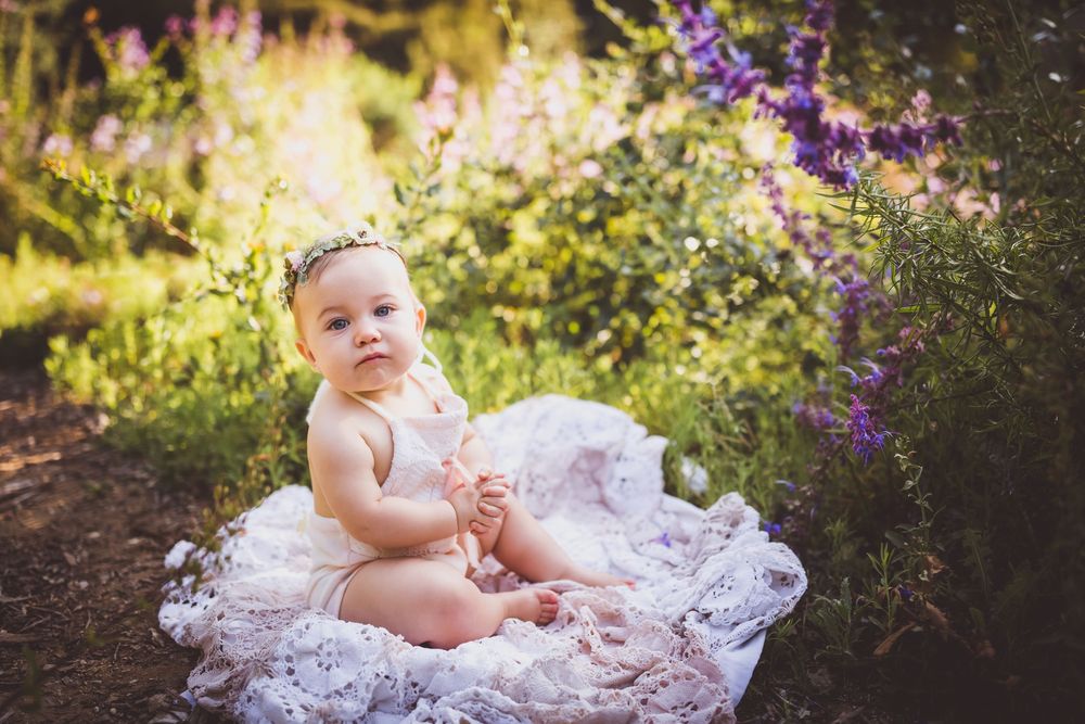 A baby girl in a white lace outfit and floral crown sits on a lace blanket surrounded by purple wildflowers outdoors.