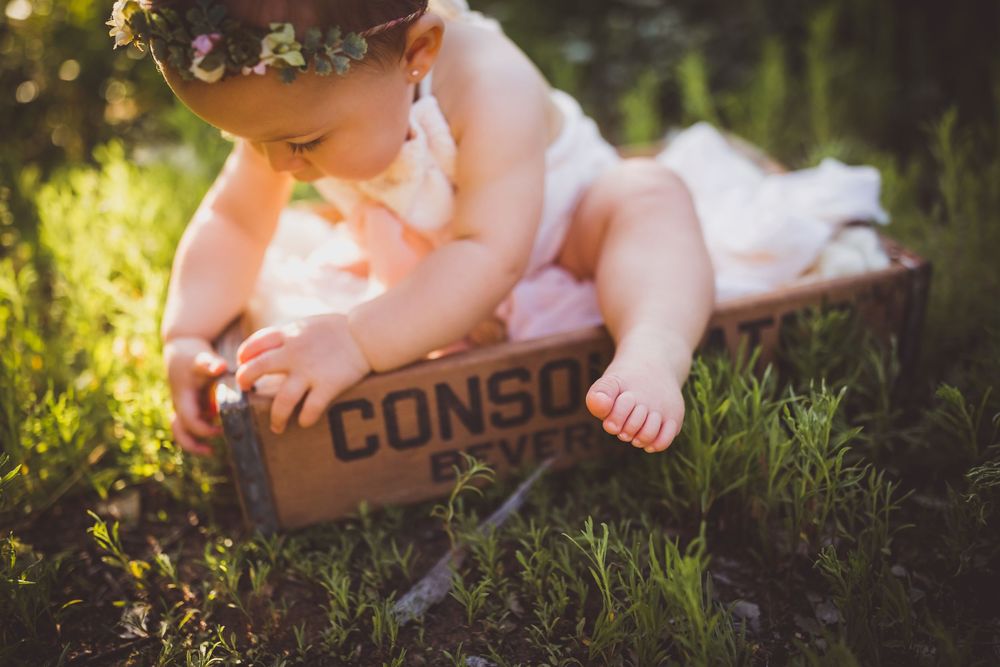 Baby girl in white dress and flower crown sitting in vintage wooden crate outdoors in green grass near yucaipa ca