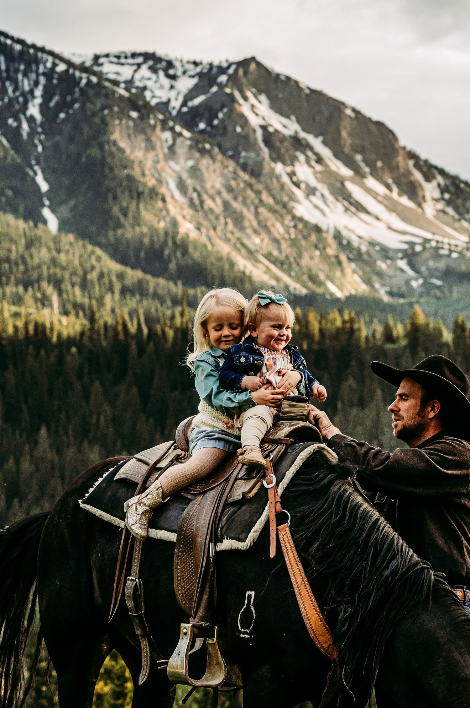 Two young girls sitting on a dark horse with a cowboy, snowy mountains and pine trees in the background.