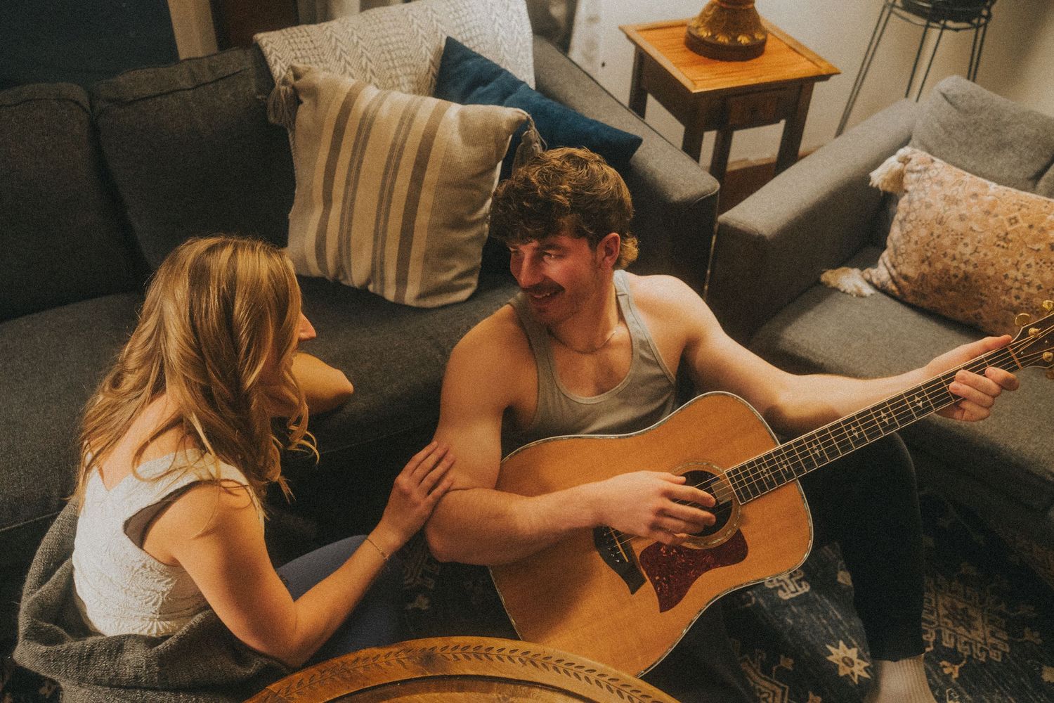 Couple sitting on floor playing acoustic guitar together in cozy living room with gray sofa and pillows.