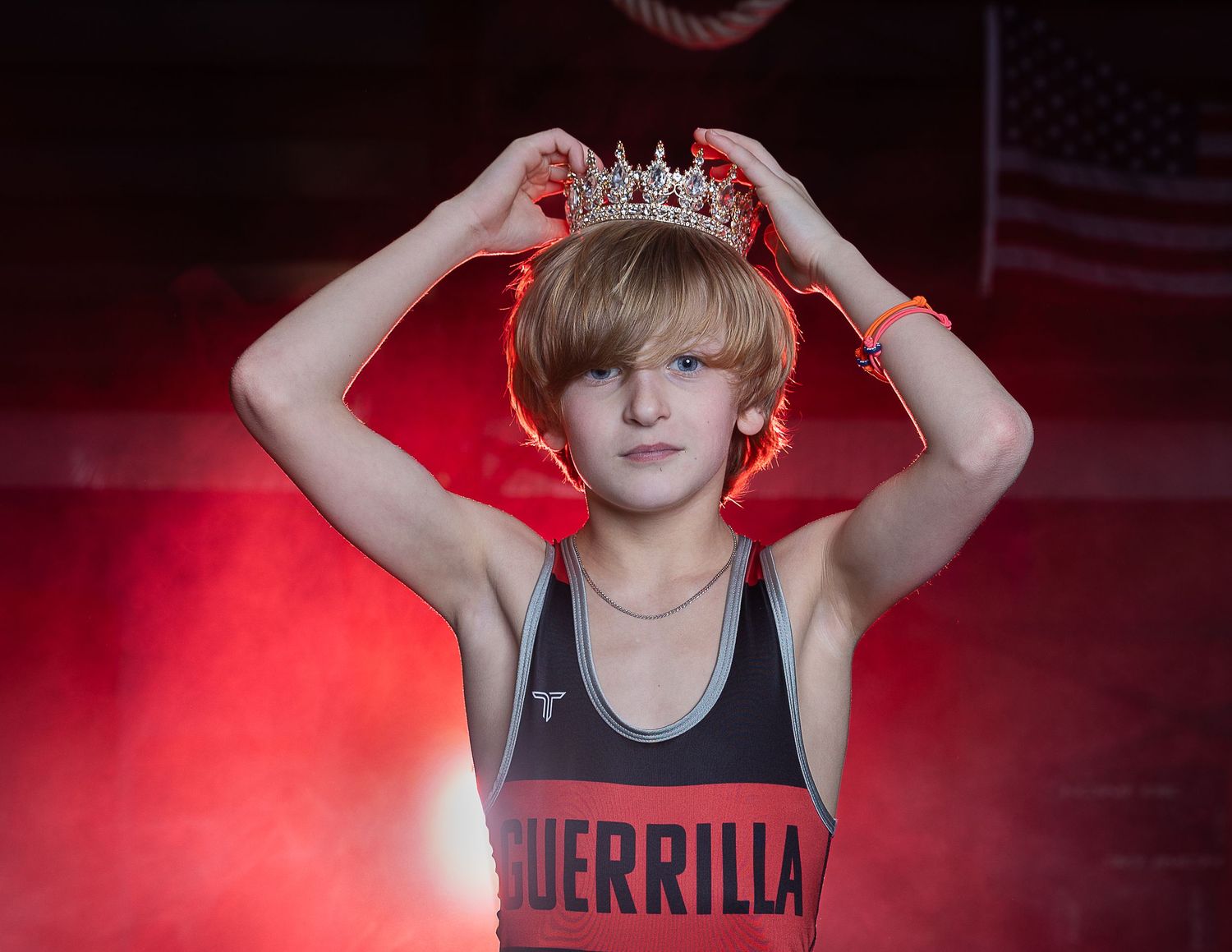 Young wrestler in Guerrilla singlet proudly placing a tiara crown on head with red dramatic lighting.