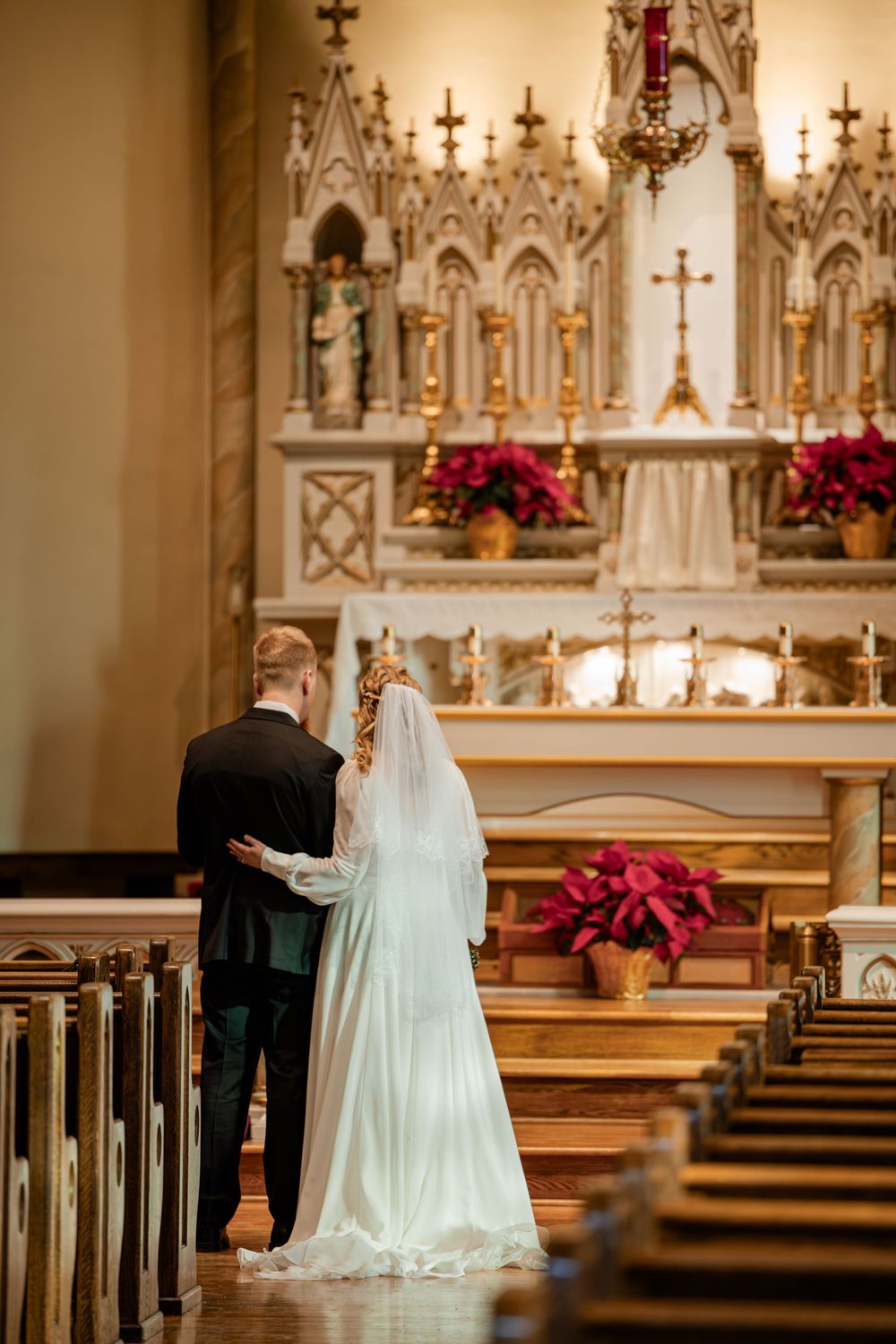 Bride and groom stand at ornate white church altar with gold crosses and red poinsettias during wedding ceremony.