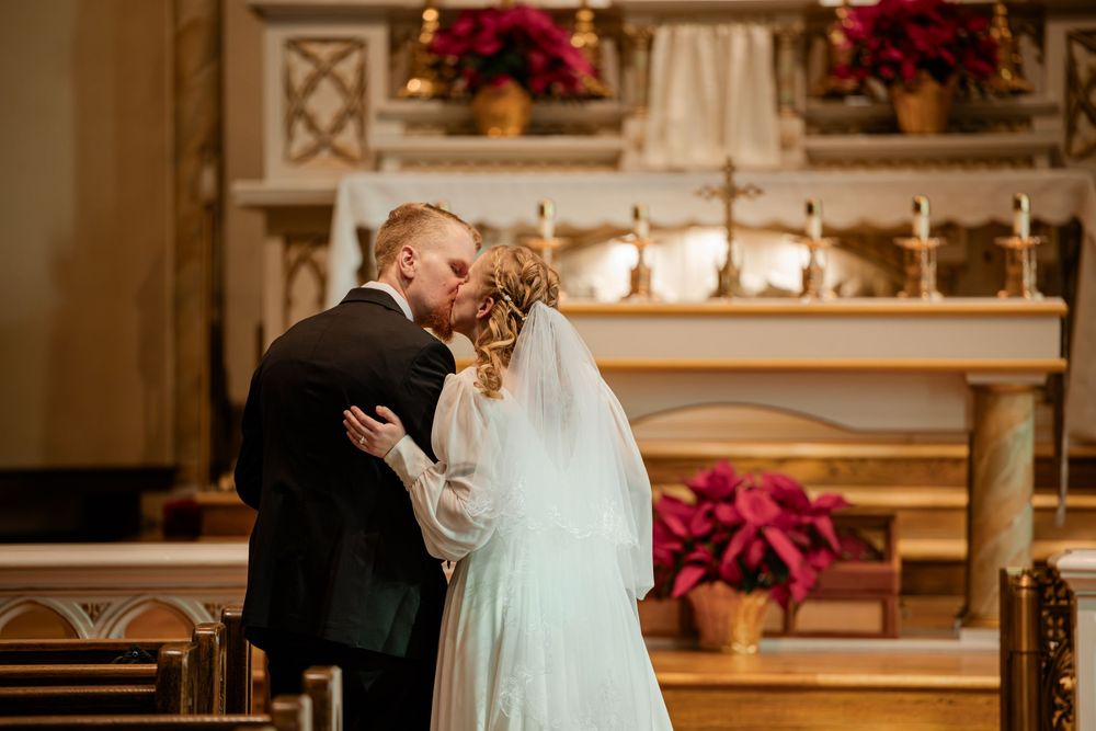 Bride and groom sharing first kiss at church altar decorated with red poinsettias during winter wedding ceremony.