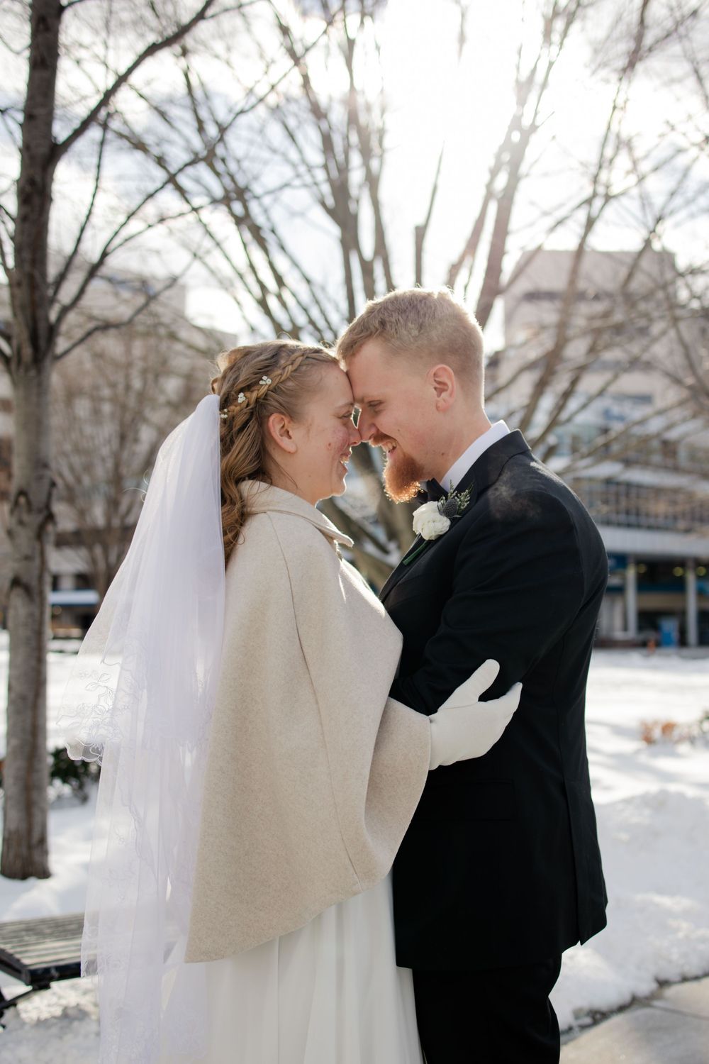 Bride and groom share a romantic winter moment outdoors, touching foreheads, dressed in wedding attire with snow-covered trees behind.