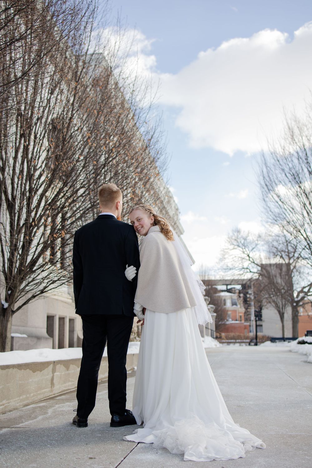 Bride in white gown and cape smiles at groom in black suit during winter outdoor wedding photo session.