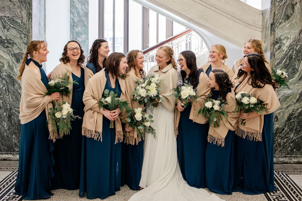 Bride with bridesmaids in teal gowns and tan shawls holding white bouquets laughing on marble staircase.