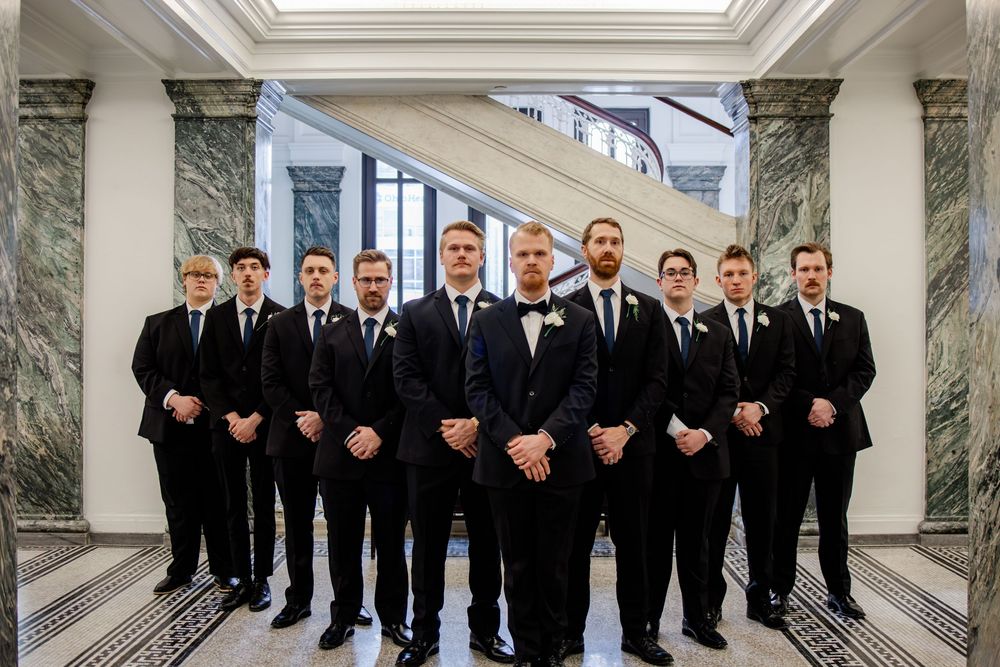 Groom and groomsmen in black tuxedos posing formally in an elegant marble hallway at a wedding venue.