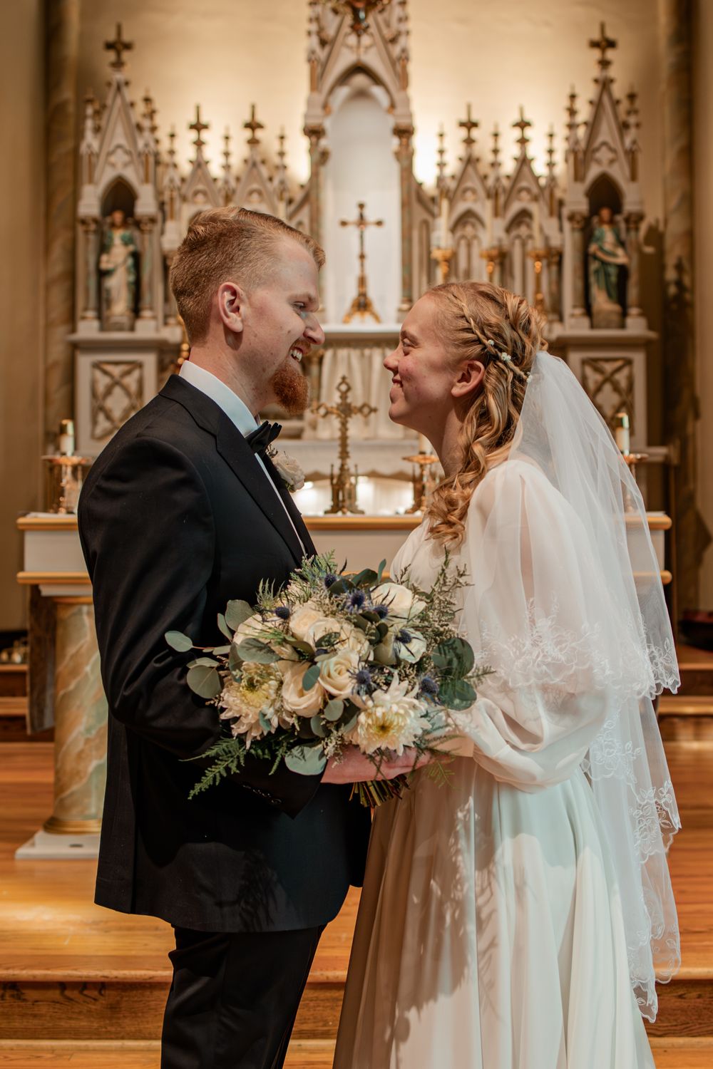 Bride and groom share a joyful moment at church altar, bride holding white floral bouquet in elegant wedding gown with veil.