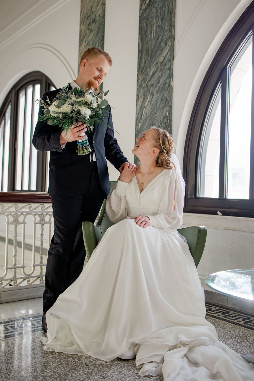 Bride in white gown seated in green chair smiles up at groom in navy suit holding wedding bouquet in elegant hall.