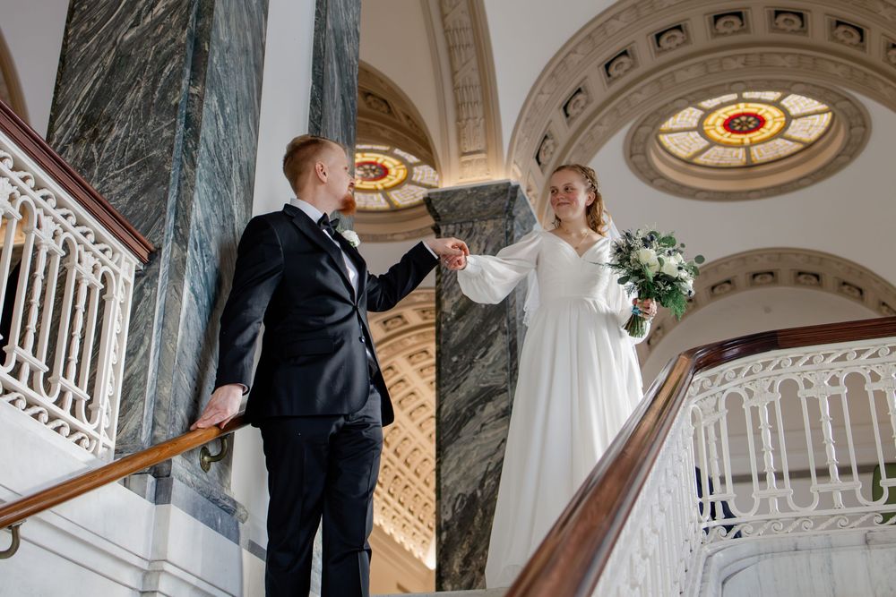 Bride and groom hold hands on elegant staircase with marble columns and ornate ceiling in a grand wedding venue.