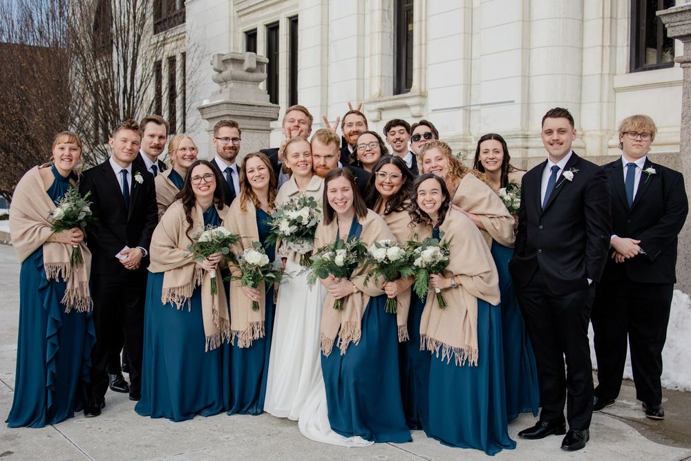 Wedding party in teal dresses and black suits posing outdoors in winter, bridesmaids wearing tan shawls holding white bouquets.