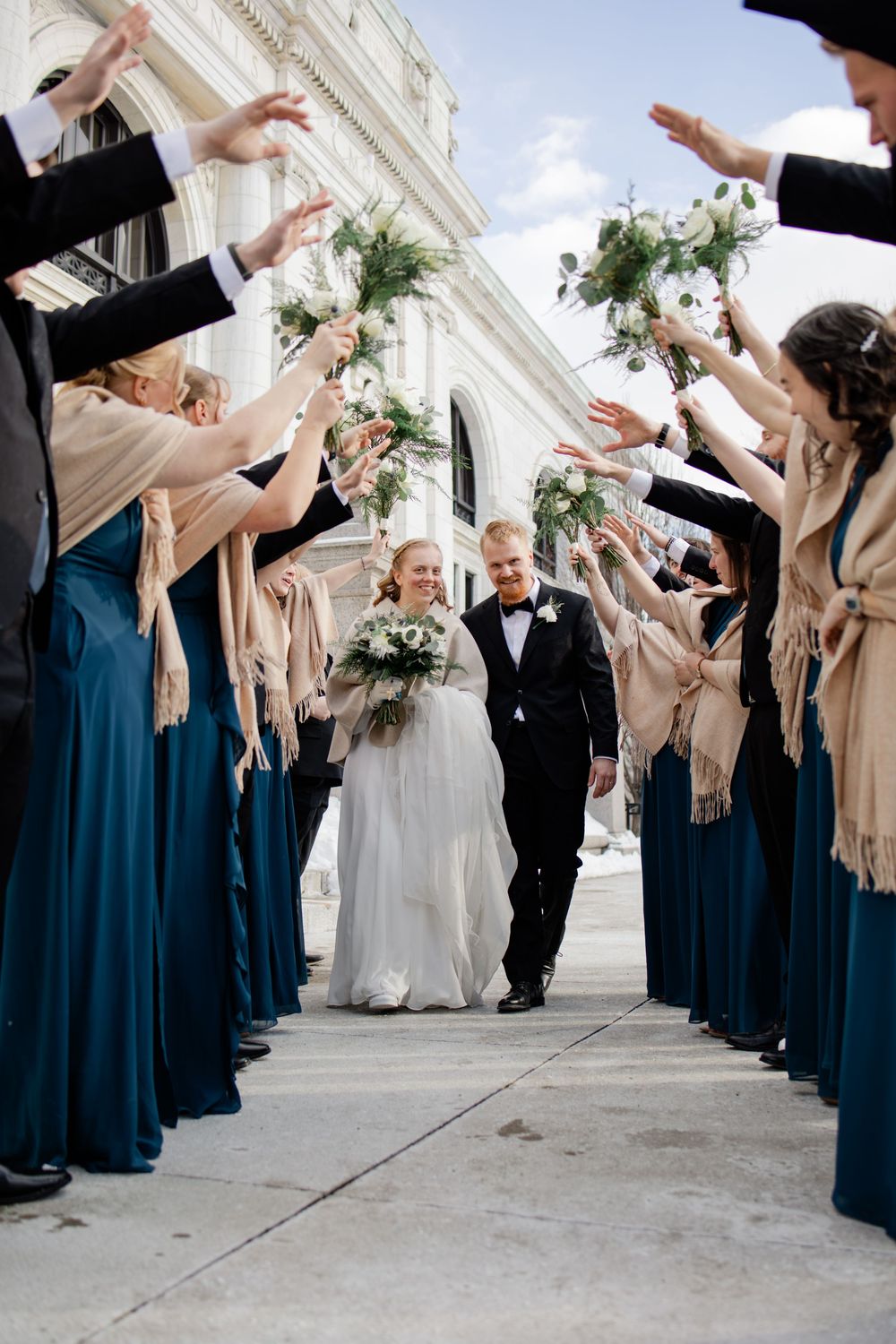 Bride and groom walk through an arch of raised hands formed by wedding party in teal gowns outdoors.