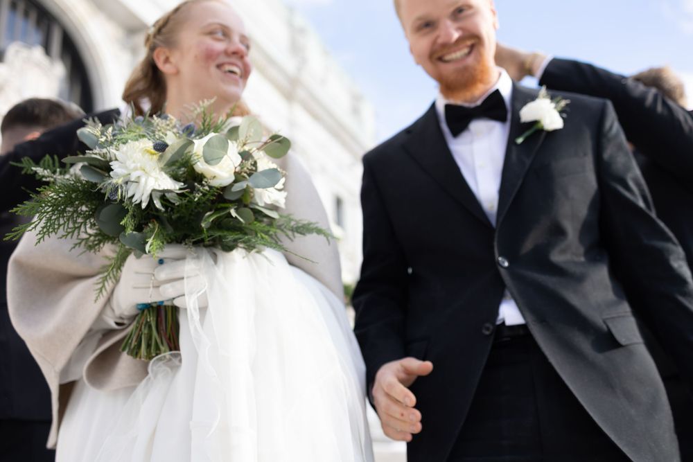 Happy bride holding white floral bouquet and groom in black tuxedo smiling outdoors on their wedding day.