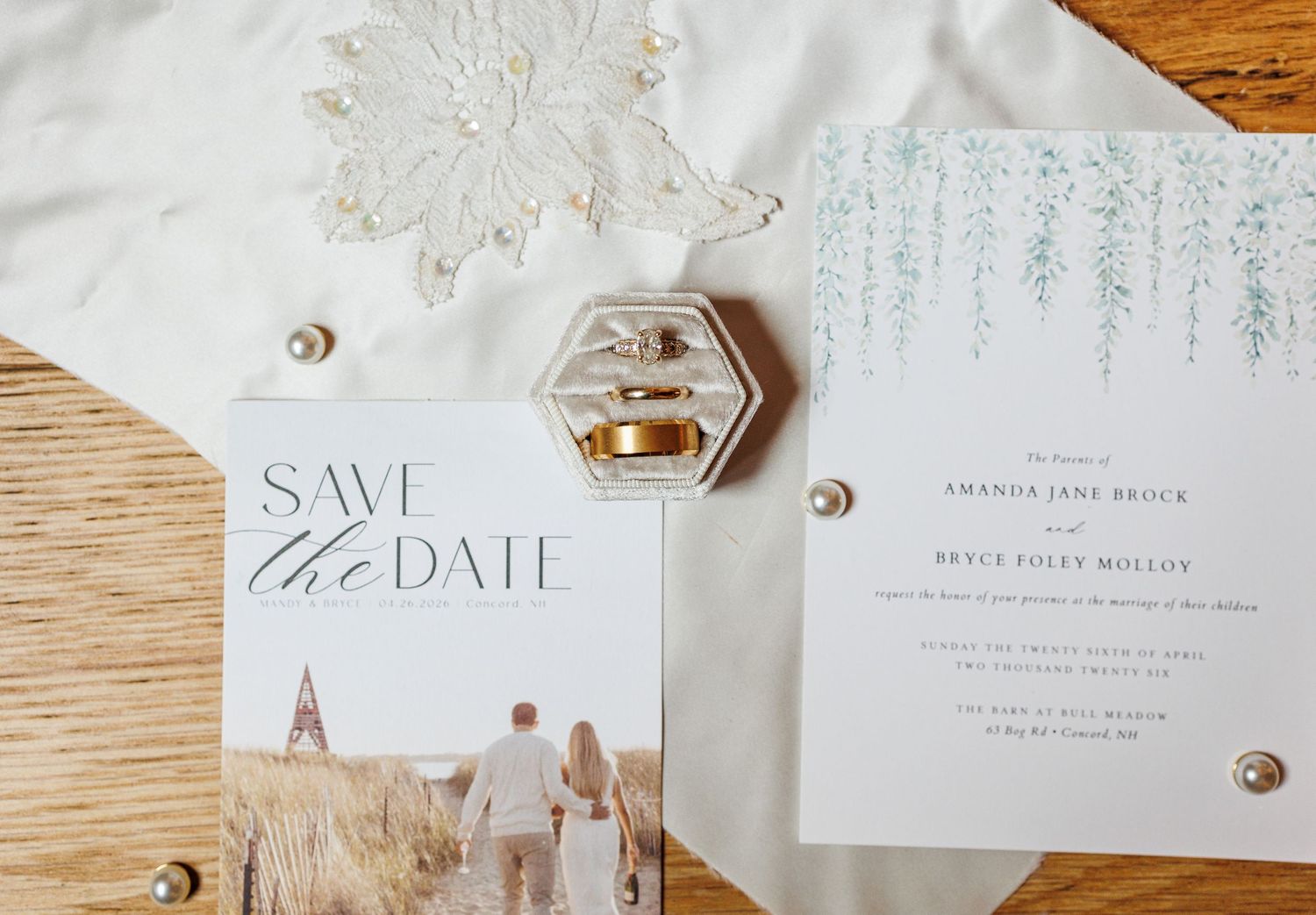 Wedding stationery flat lay with save the date card, formal invitation, rings in acrylic box, and lace veil on wood.