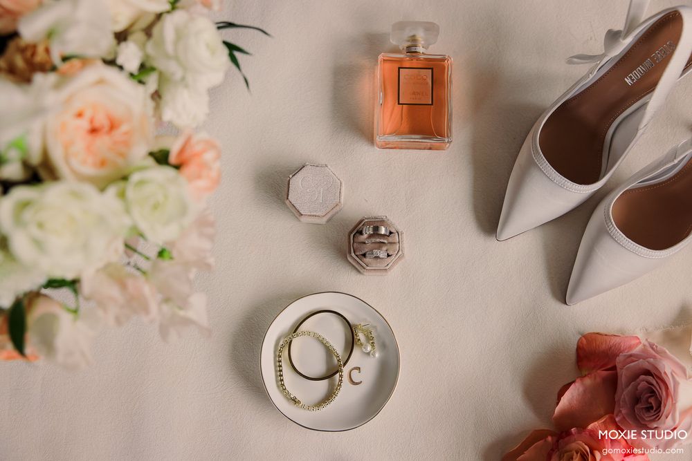 Bridal flat lay with perfume, white heels, jewelry, and flowers on a neutral background.