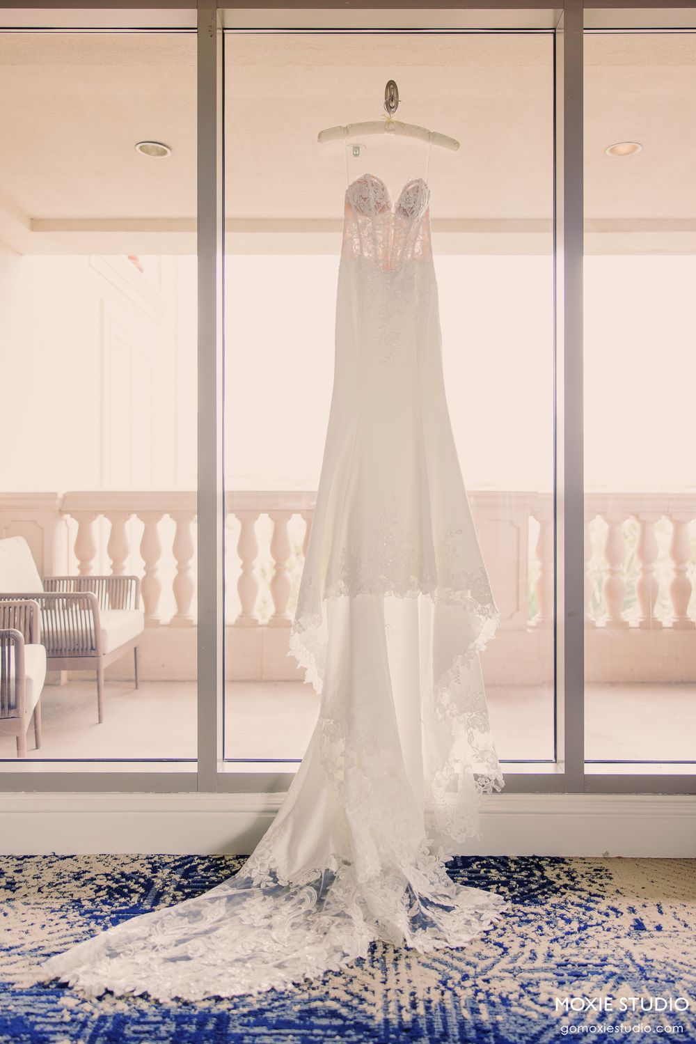 Elegant strapless lace wedding gown with long train hanging in a sunlit window with balcony view.