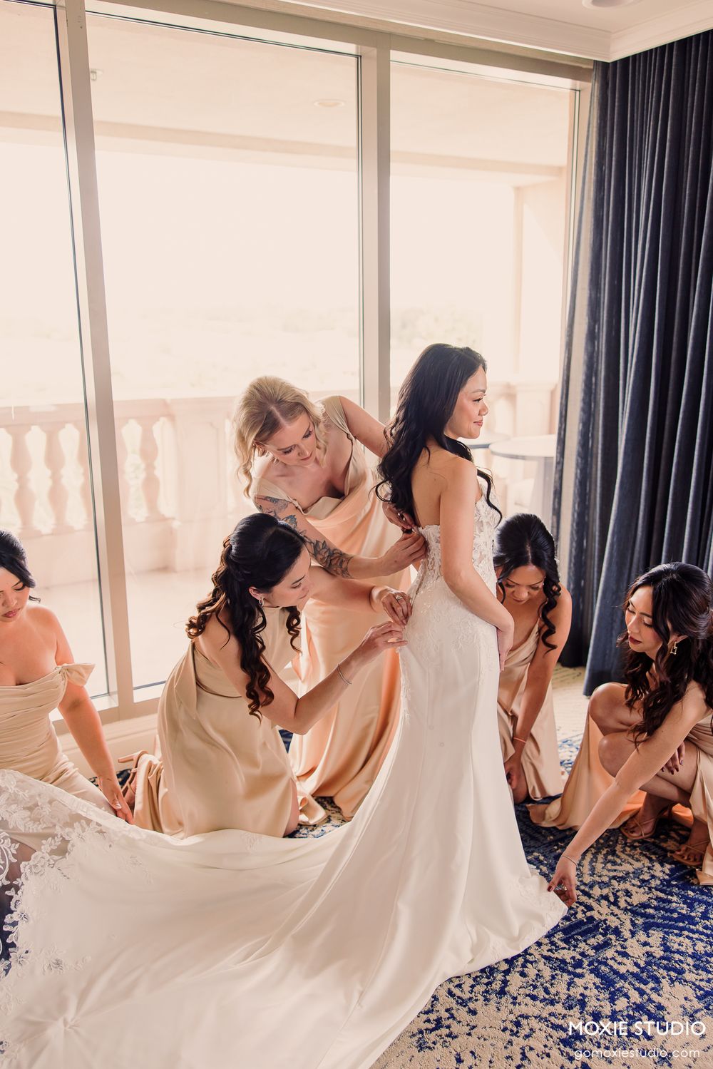 Bridesmaids in champagne gowns help bride into white wedding dress near sunlit balcony windows.