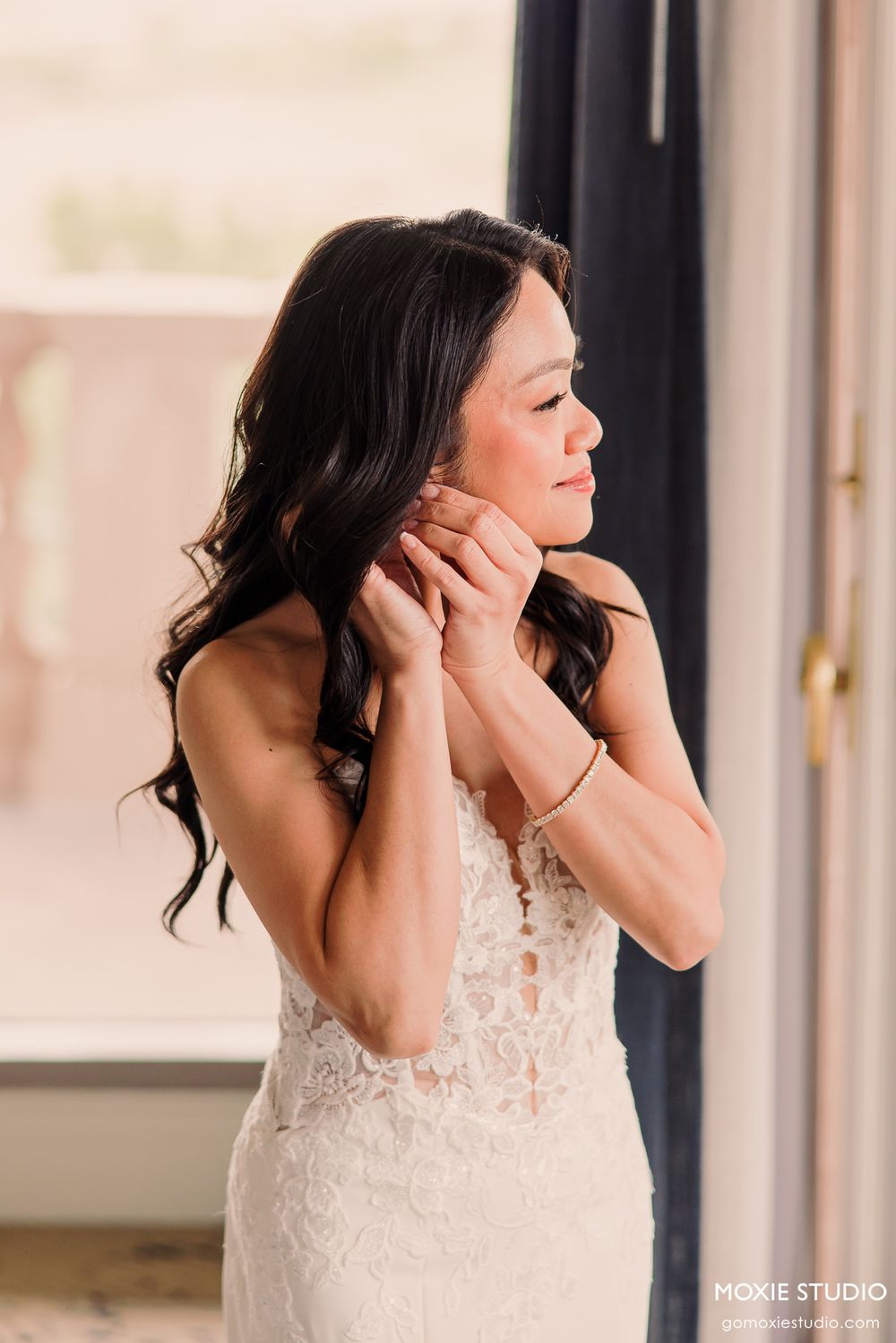 Bride in lace wedding gown adjusting earrings near window, dark wavy hair, elegant bridal getting ready photo.