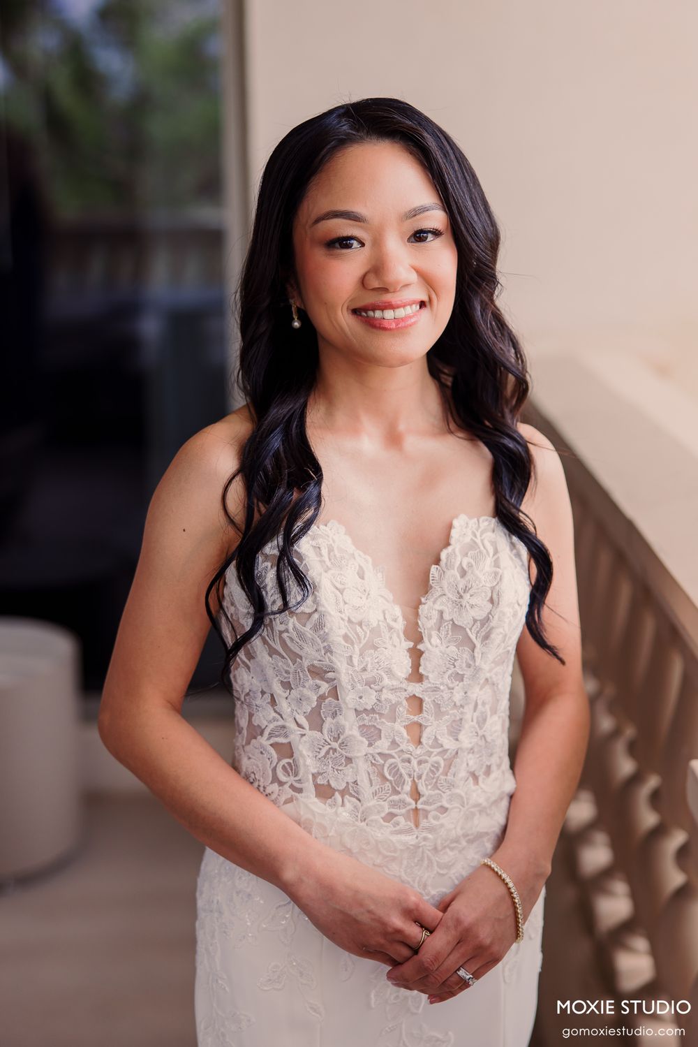 Smiling bride in lace wedding gown poses elegantly on a balcony, showcasing her beautiful bridal look.