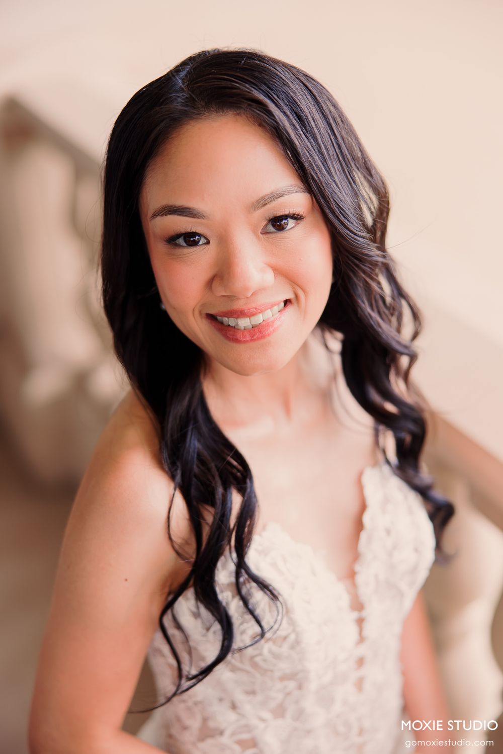 Smiling Asian bride in white lace gown with elegant curled hair and natural makeup, professional wedding portrait.