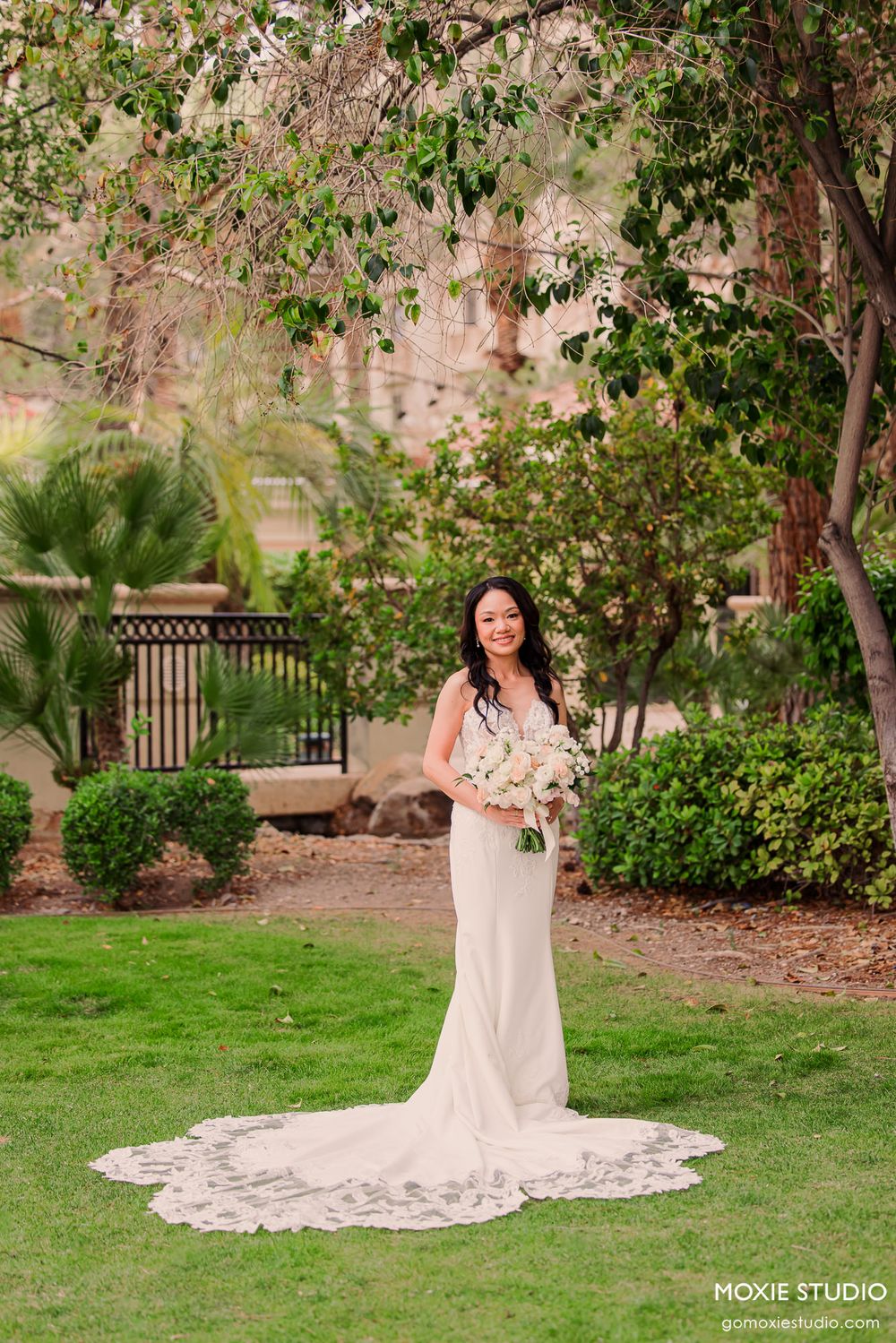 Bride in elegant white gown with lace train holds bouquet while posing in lush garden setting.