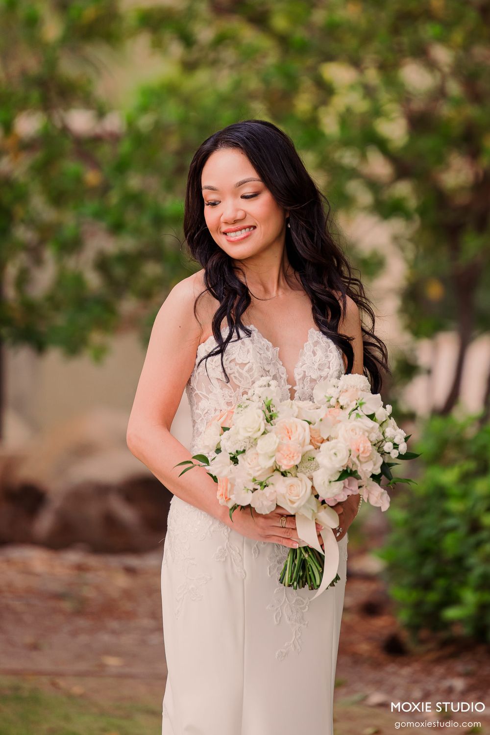 Smiling bride in lace gown holding white and blush rose bouquet, standing outdoors with greenery background.