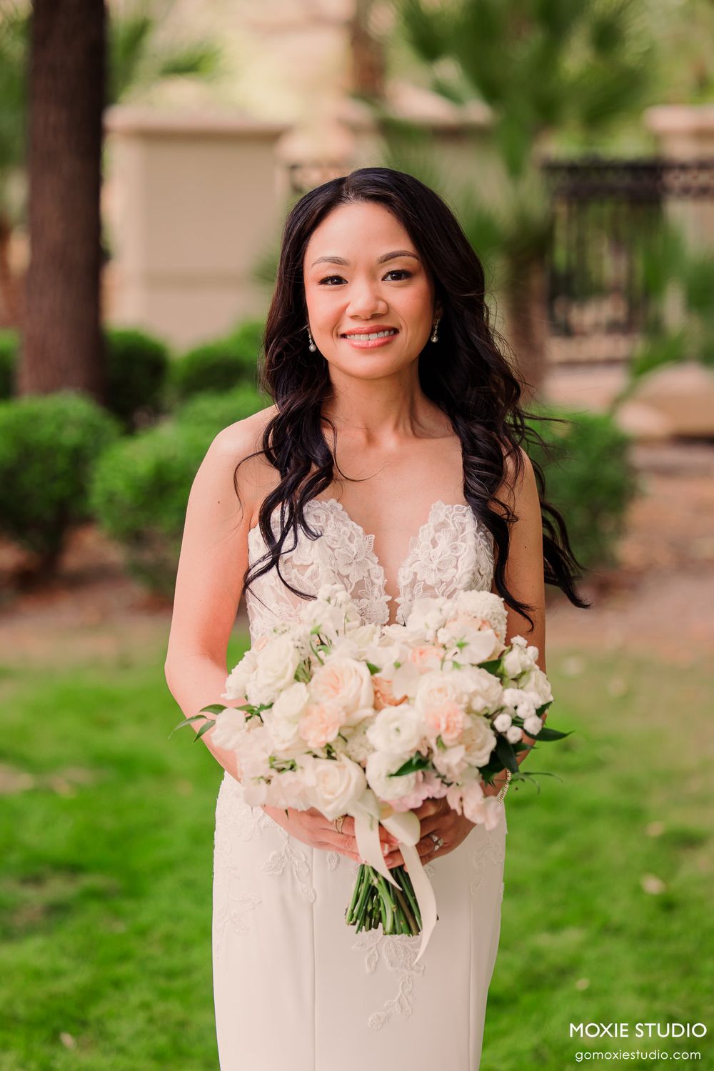 Smiling bride in lace wedding gown holding elegant white and blush bouquet outdoors.