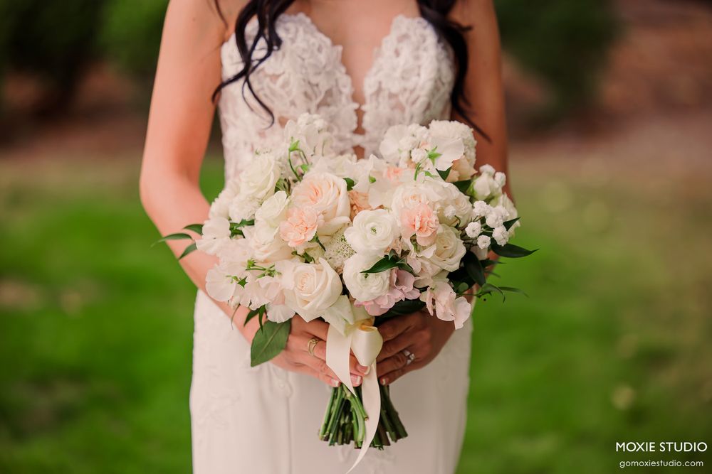 Bride in lace wedding gown holding elegant blush pink and white floral bouquet with roses and greenery outdoors.