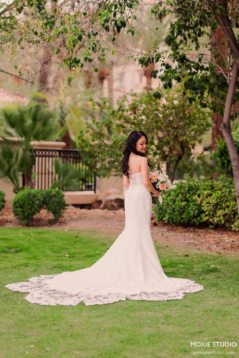 Bride in elegant strapless white gown with lace train holds bouquet in lush garden setting.