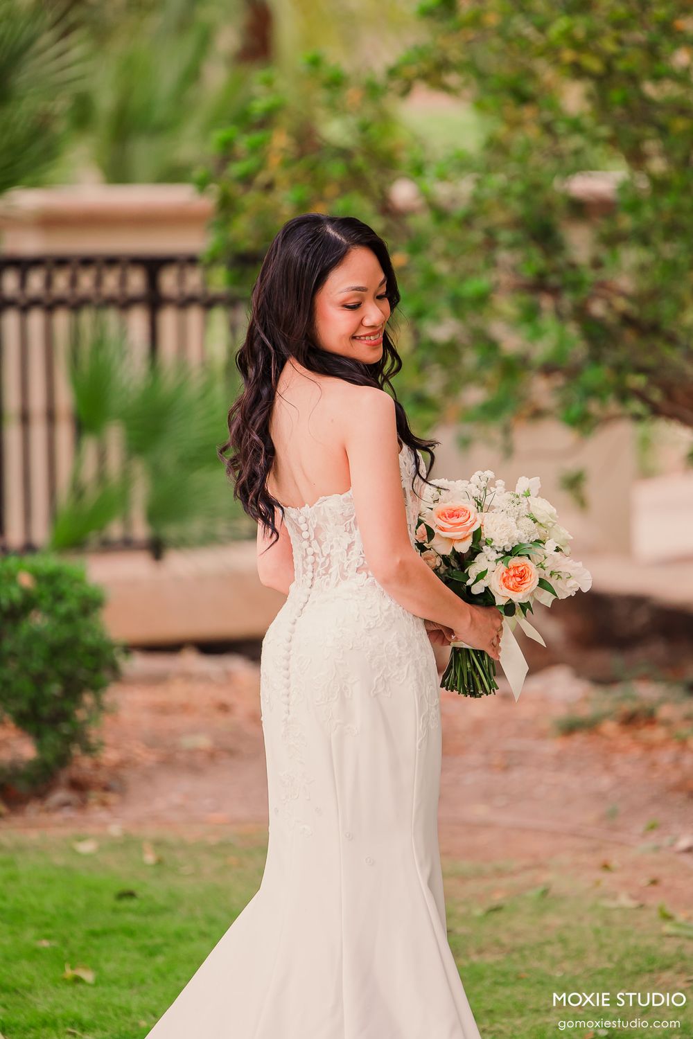 Bride in a backless lace wedding gown holding a peach and white rose bouquet, smiling over her shoulder outdoors.