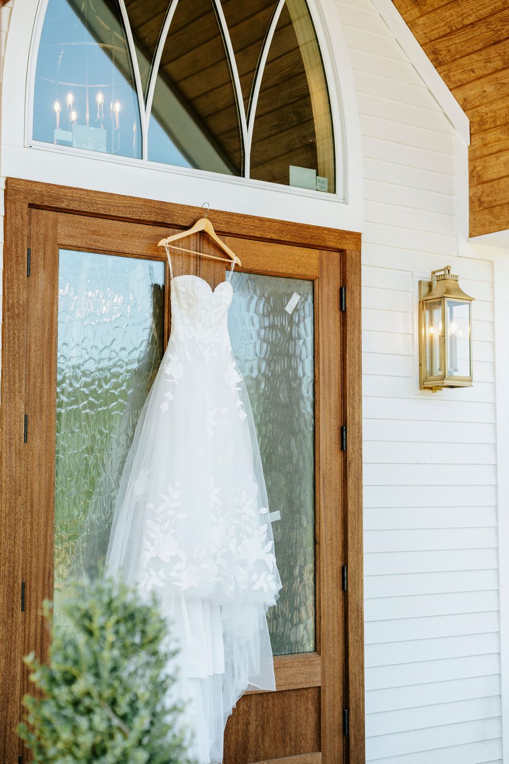 A white lace wedding dress hanging on a wooden door of a charming white chapel with a brass wall lantern.
