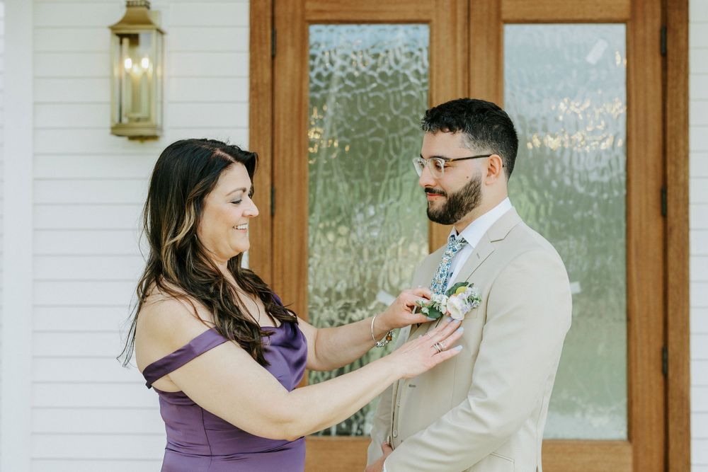 A woman in a purple dress pins a boutonniere on a groom in a tan suit outside a white building.