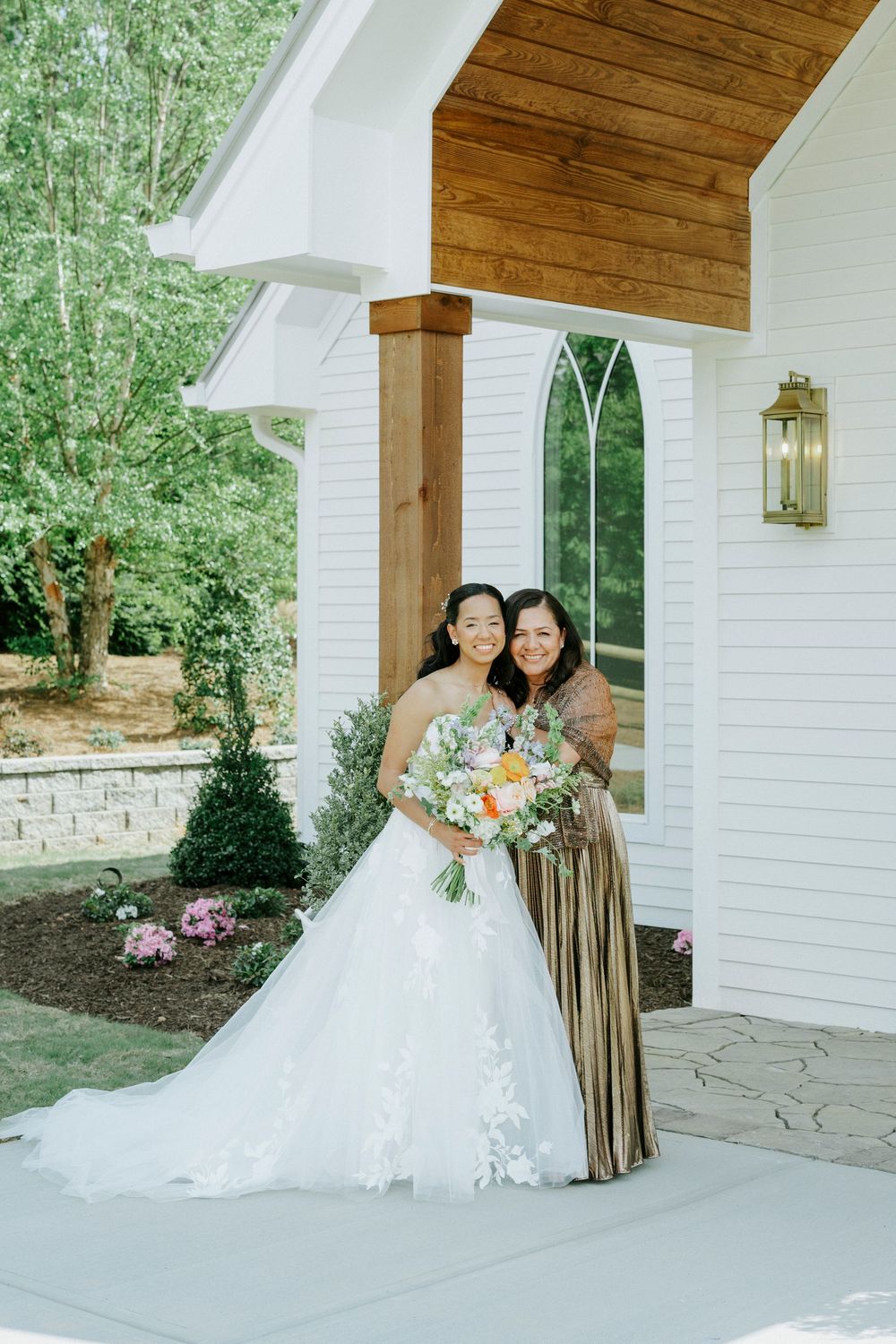 Bride in white lace gown holds colorful bouquet beside woman in gold dress outside white chapel.