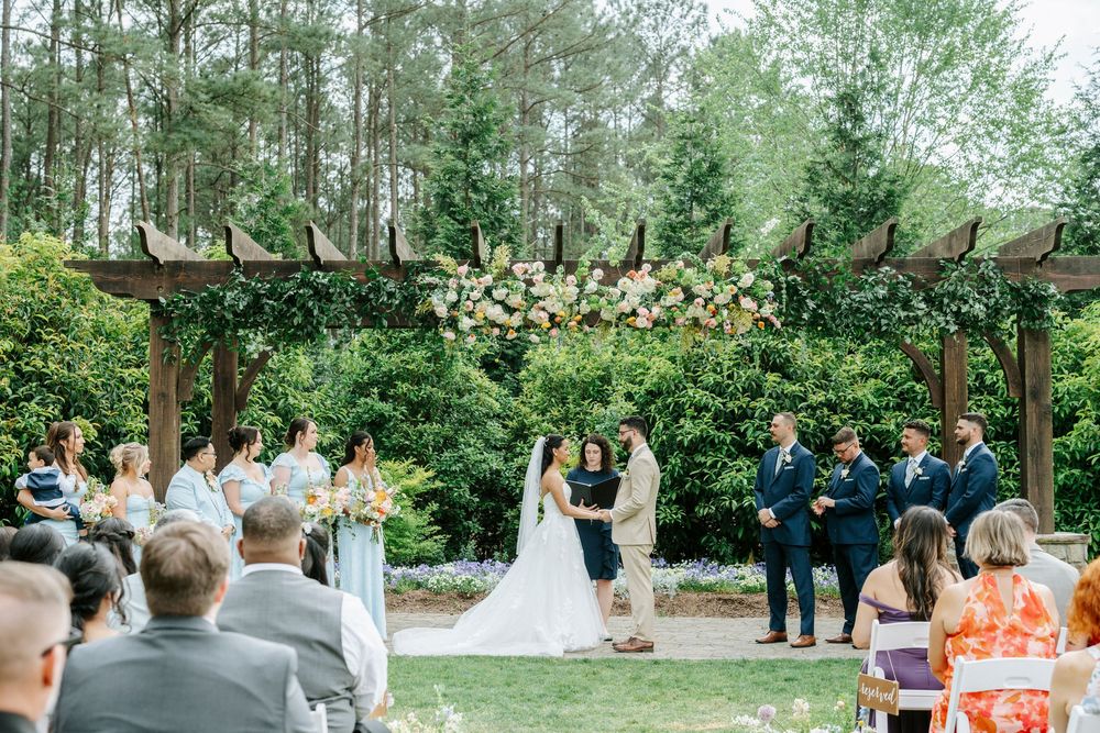Outdoor wedding ceremony under a floral-adorned pergola surrounded by lush greenery, with guests seated on a sunny day.