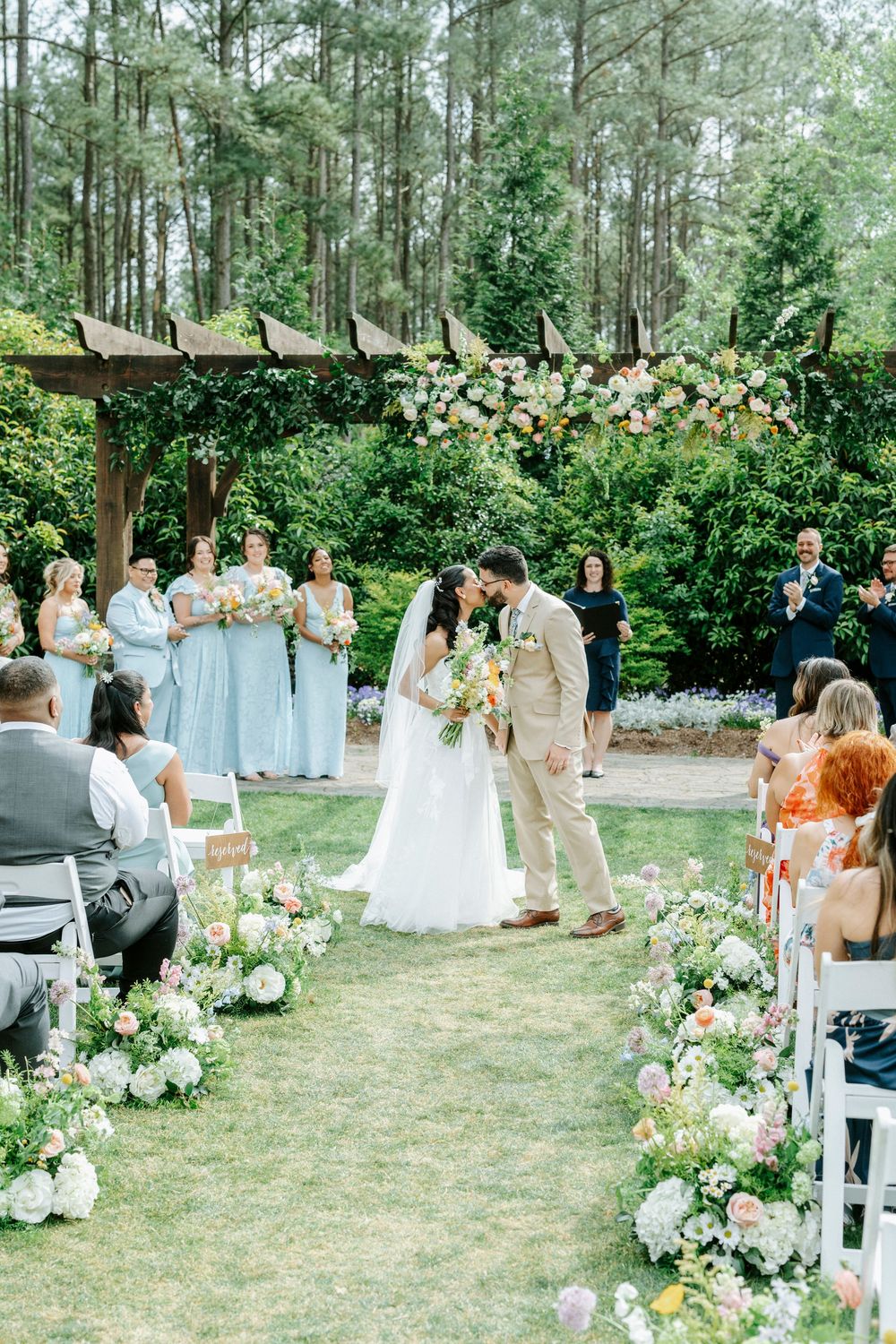 Bride and groom share first kiss at outdoor garden wedding ceremony with floral aisle and bridesmaids in light blue dresses.