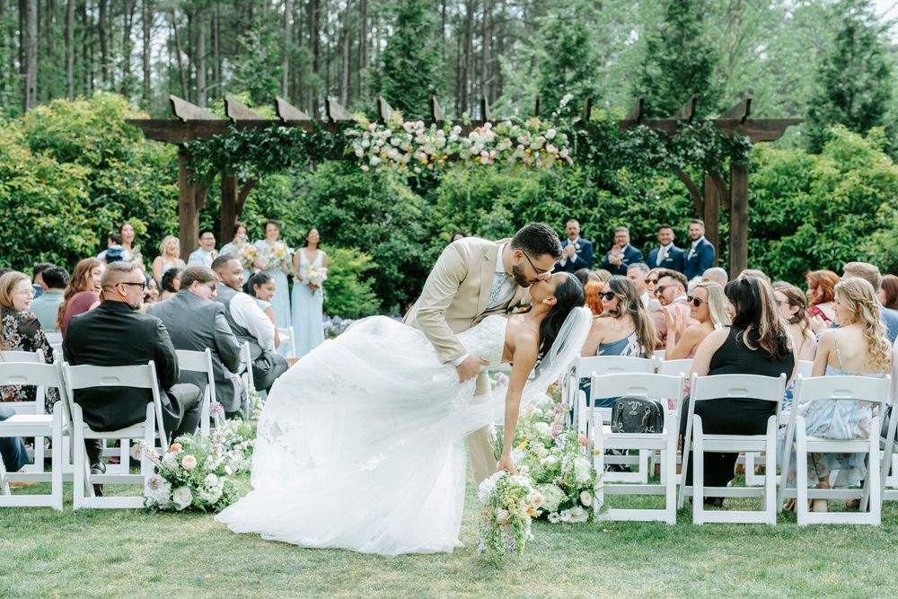 Bride and groom share a romantic dip kiss during outdoor wedding ceremony surrounded by guests and floral arch.