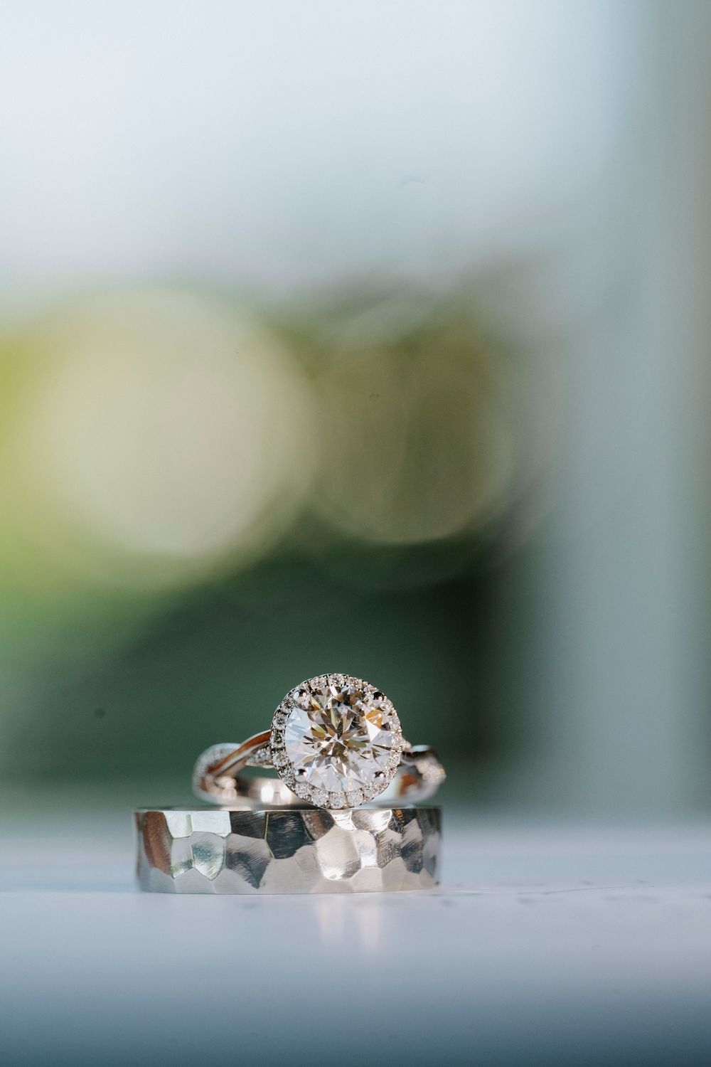 Elegant round diamond halo engagement ring resting on a hammered silver wedding band on a white surface.