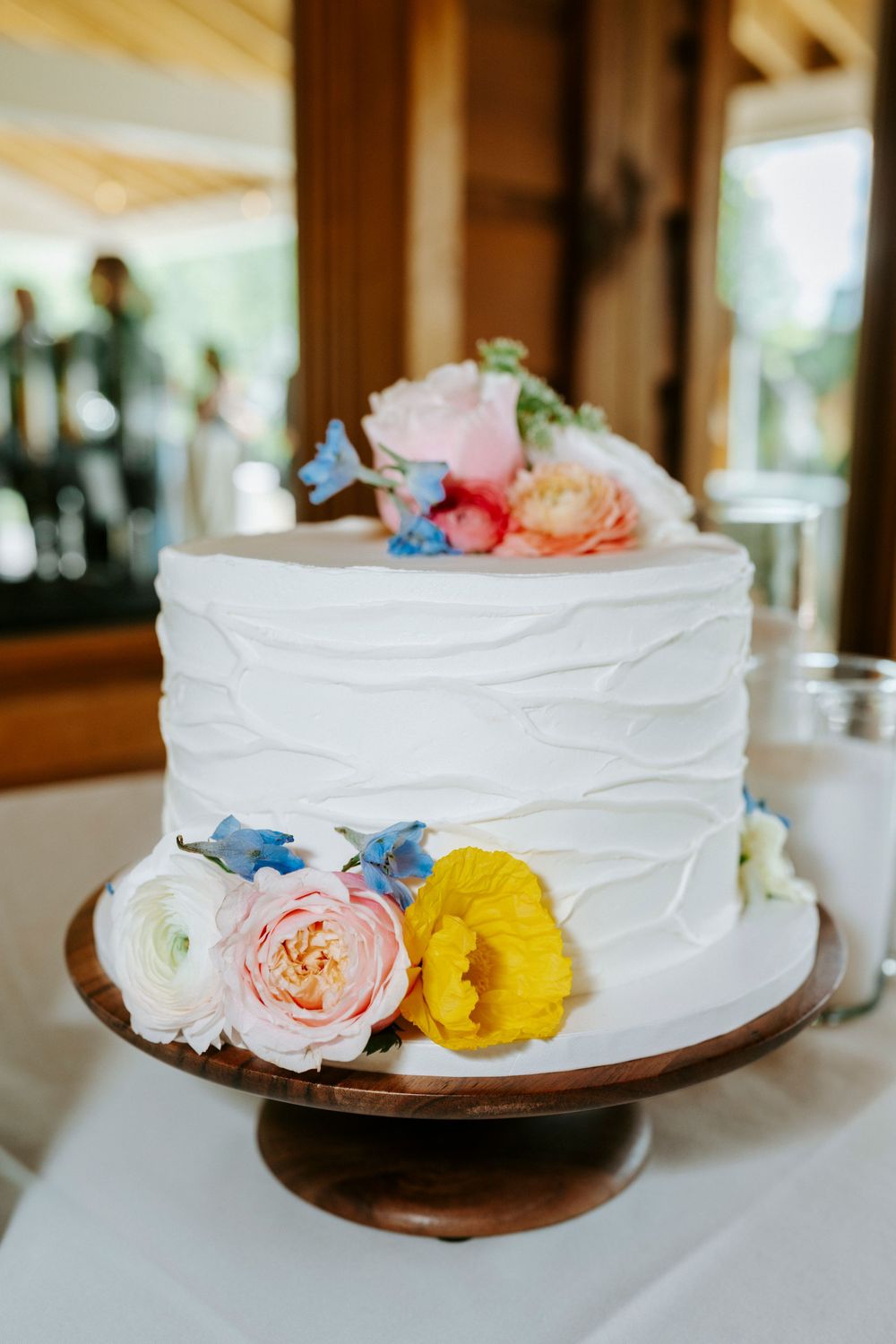 Single-tier white textured wedding cake decorated with colorful pink, yellow, and blue flowers on a wooden cake stand.