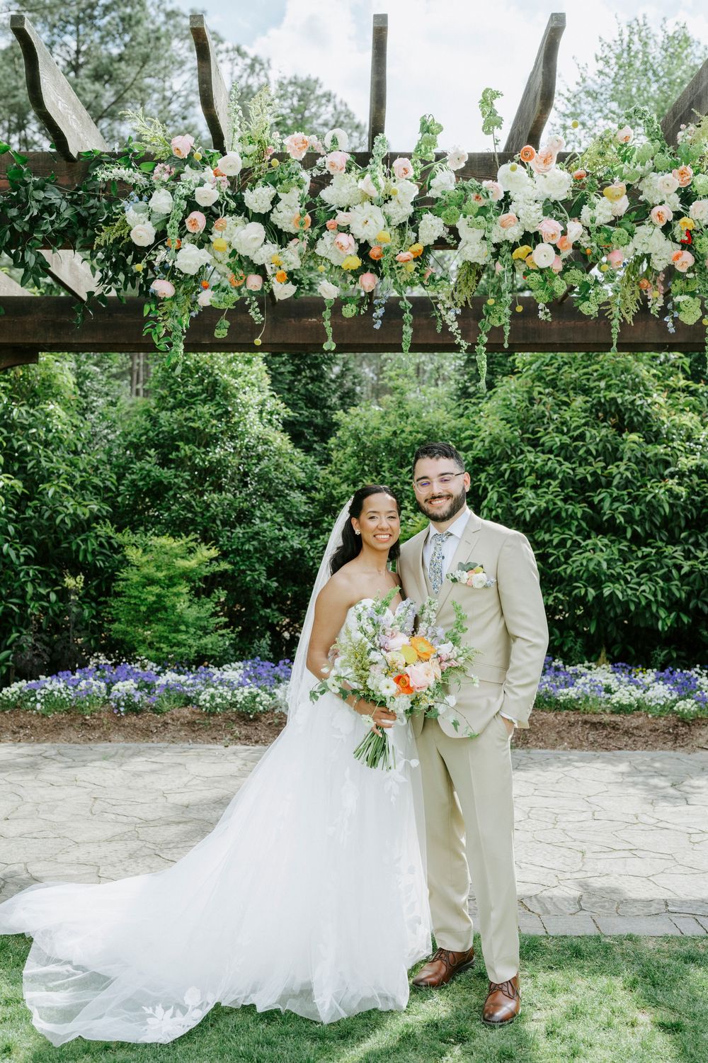 Bride and groom pose under a floral pergola with colorful blooms, lush greenery, and a stone patio at an outdoor wedding.