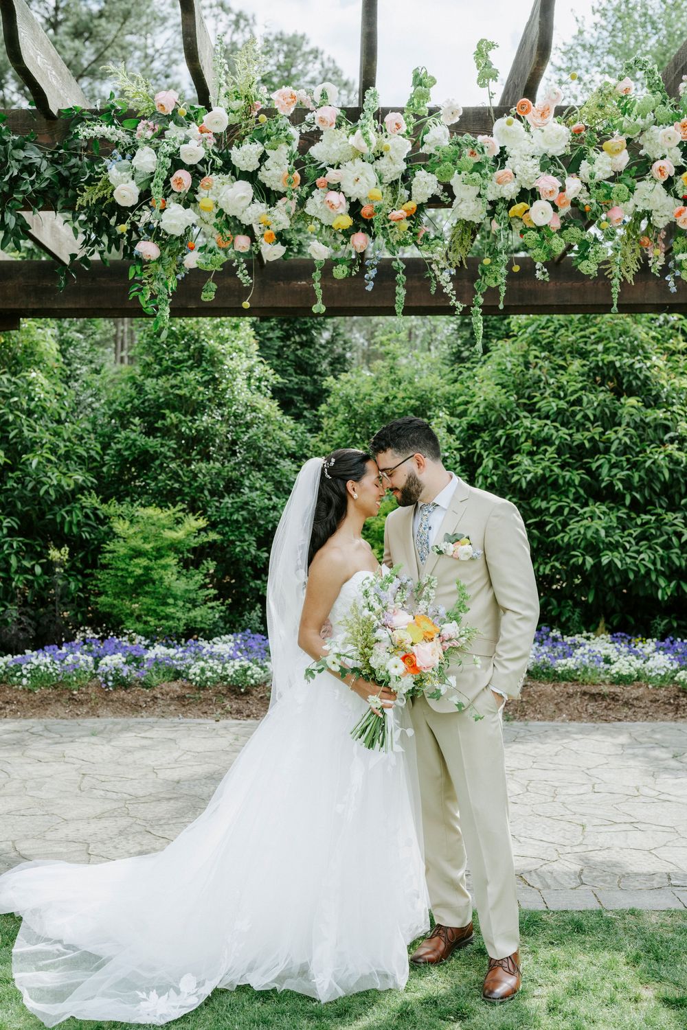 Bride and groom embrace beneath a colorful floral pergola, bride holding a pastel bouquet in a garden setting.