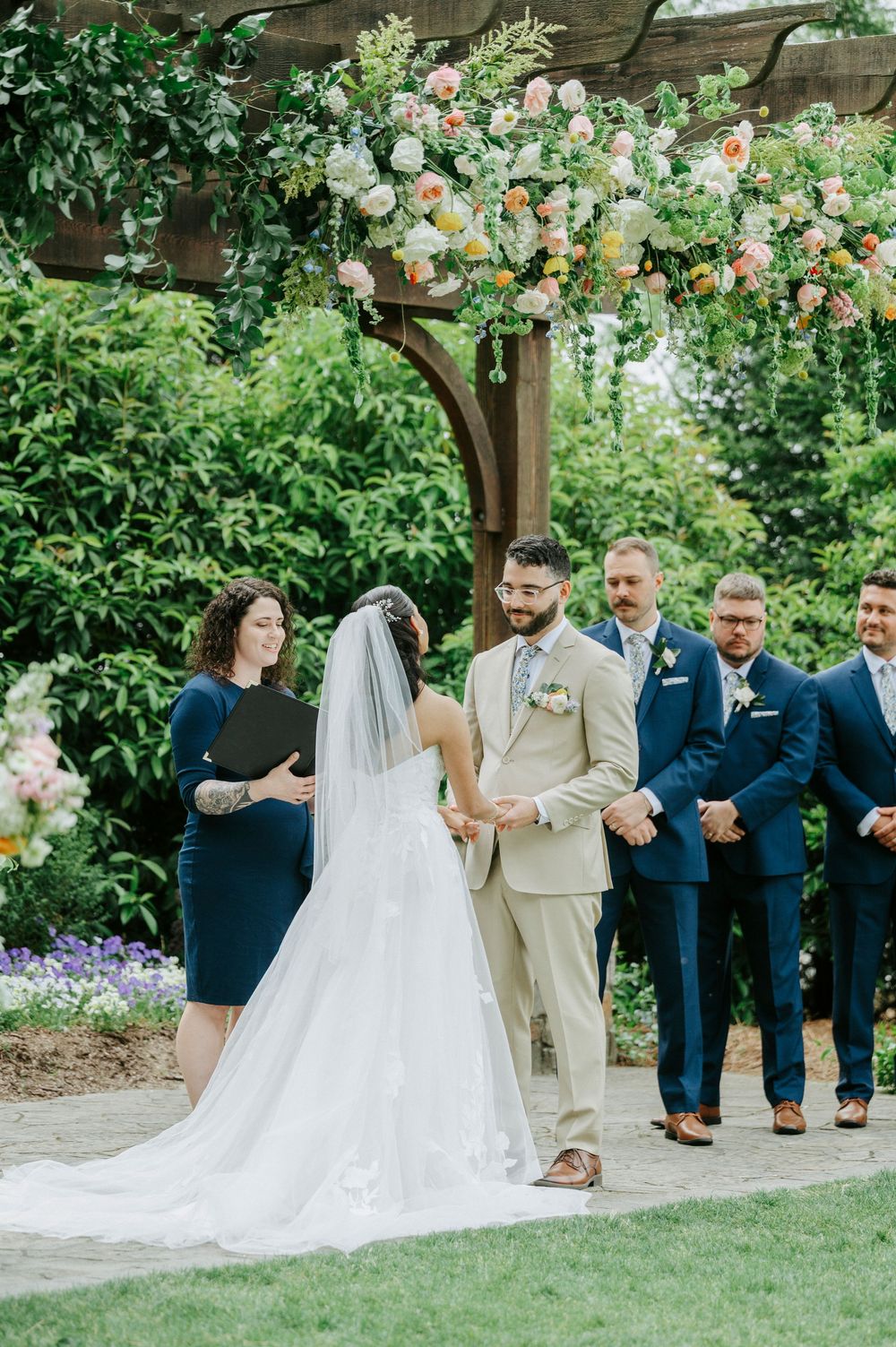 Bride and groom exchange vows under a floral arch at an outdoor garden wedding ceremony.