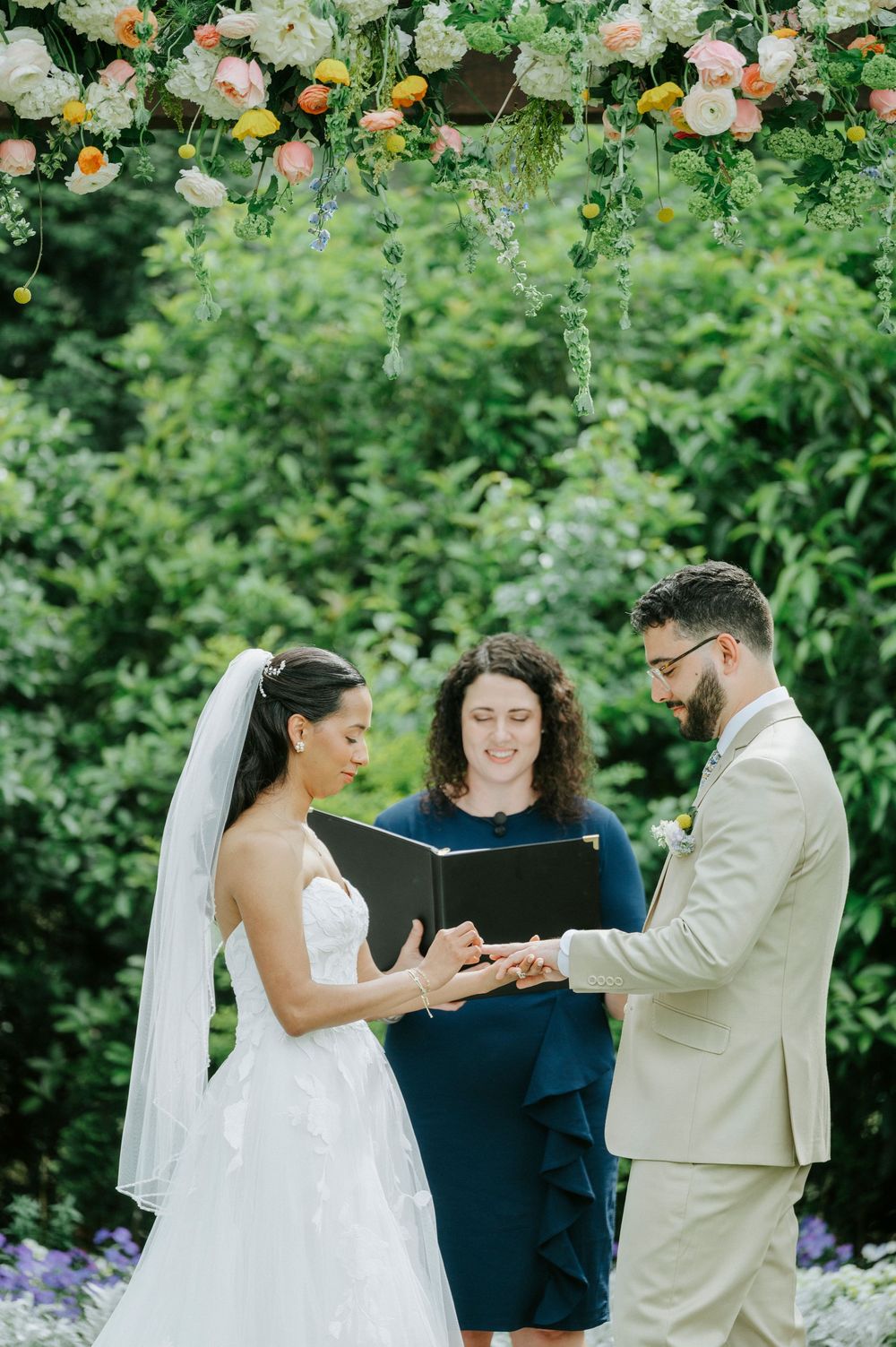 Bride places ring on grooms finger during outdoor wedding ceremony beneath floral arch with officiant.