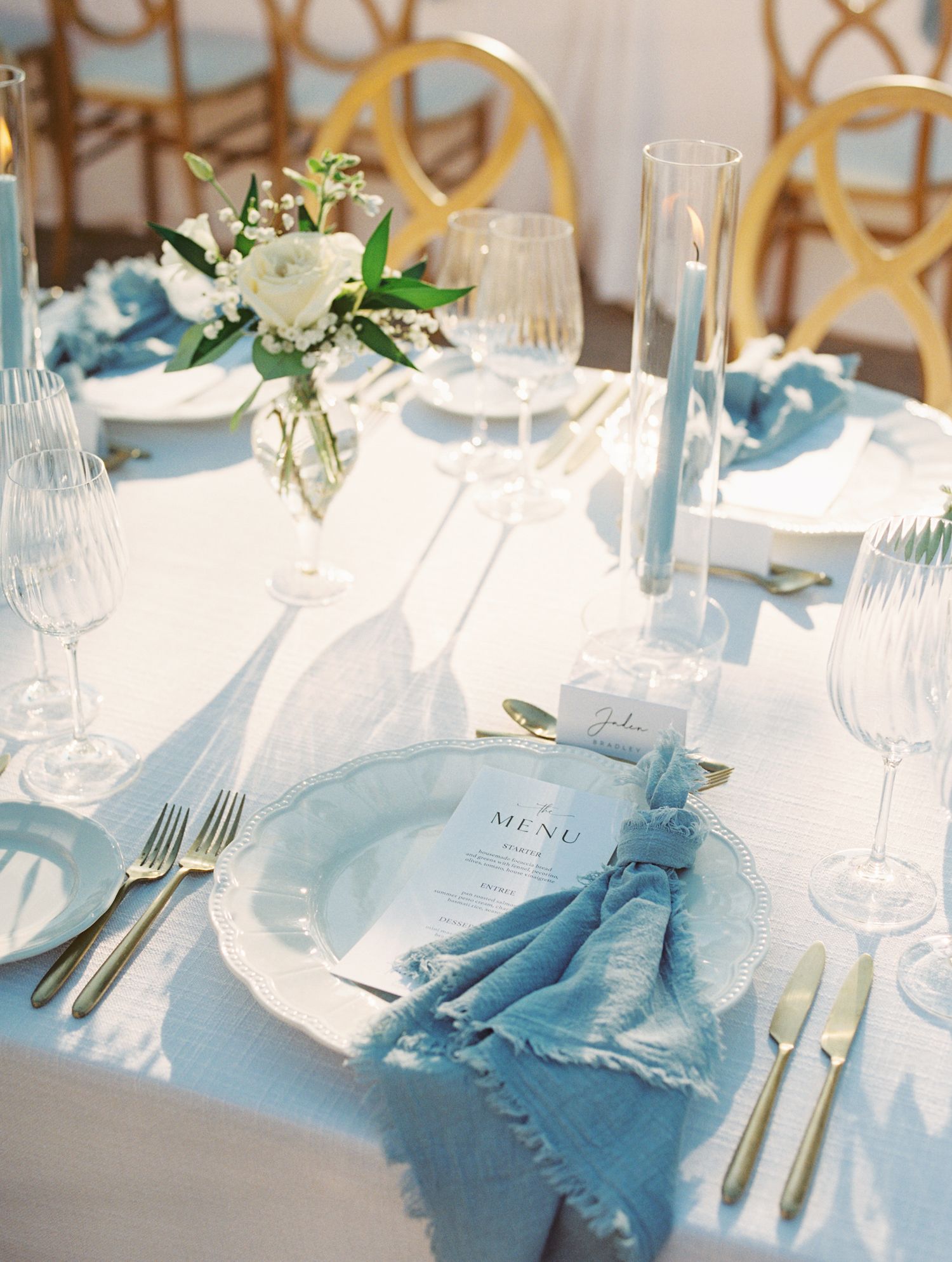 Elegant blue and white wedding tablescape with gold chairs, floral centerpiece, crystal glassware, and blue taper candles.