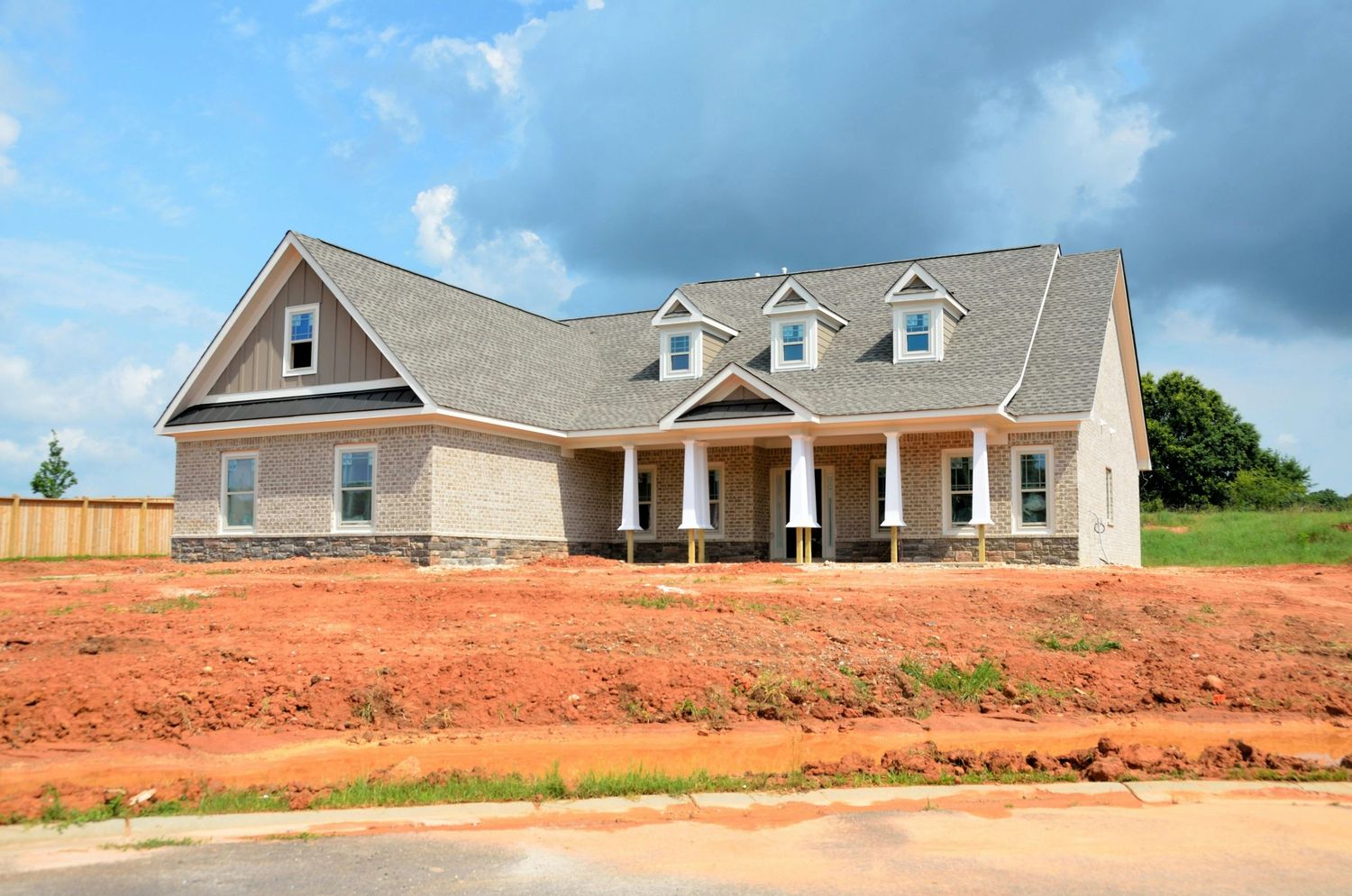 New construction craftsman-style home with dormers and covered porch surrounded by red clay soil under cloudy skies.