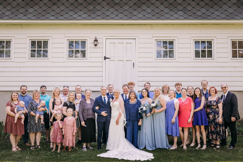 Wedding couple poses with large family group outside white barn venue on sunny day.