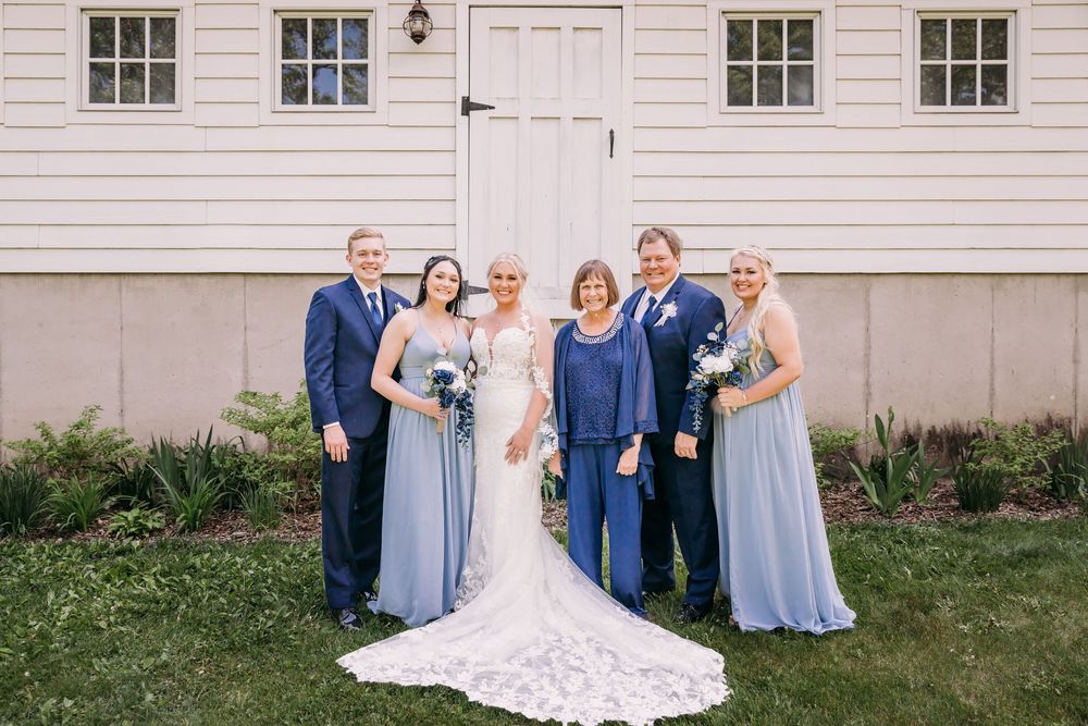 Wedding party in dusty blue dresses poses with bride and groom before white barn.